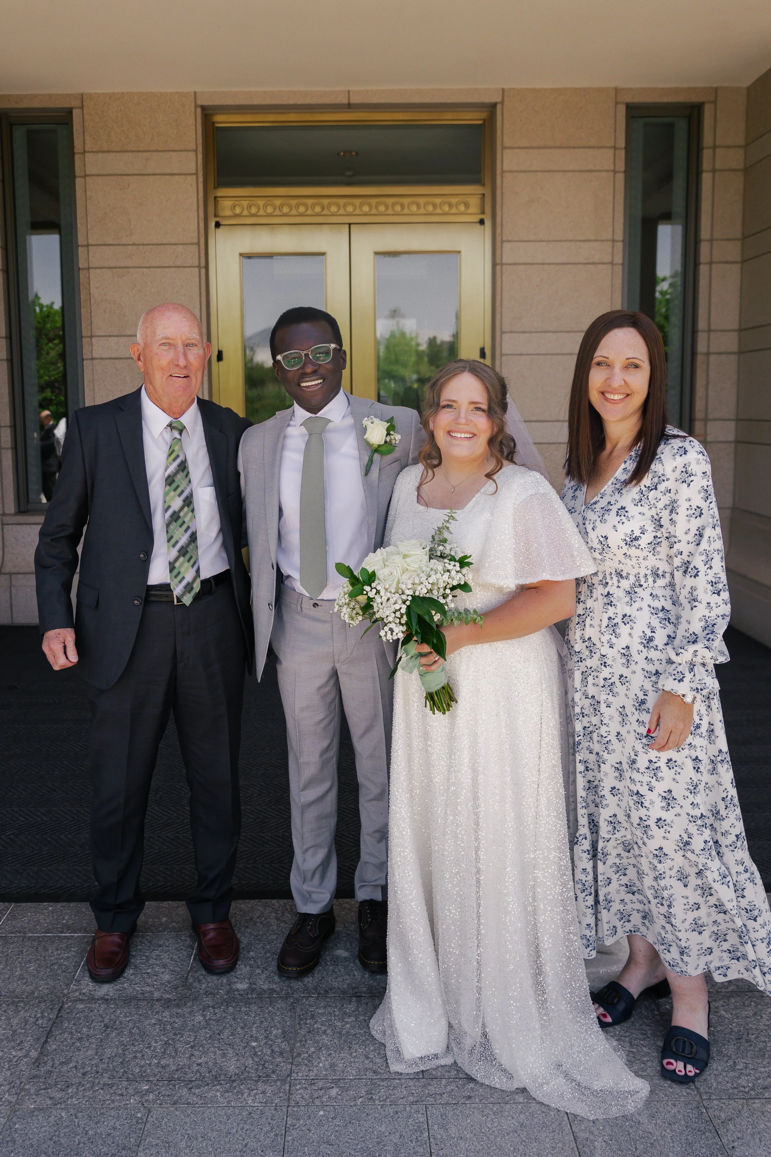Group of four people standing outside a building, celebrating a wedding. The bride is holding a bouquet of white flowers, wearing a white wedding dress, and smiling. The groom is in a light gray suit with a boutonniere. An older man is in a dark suit