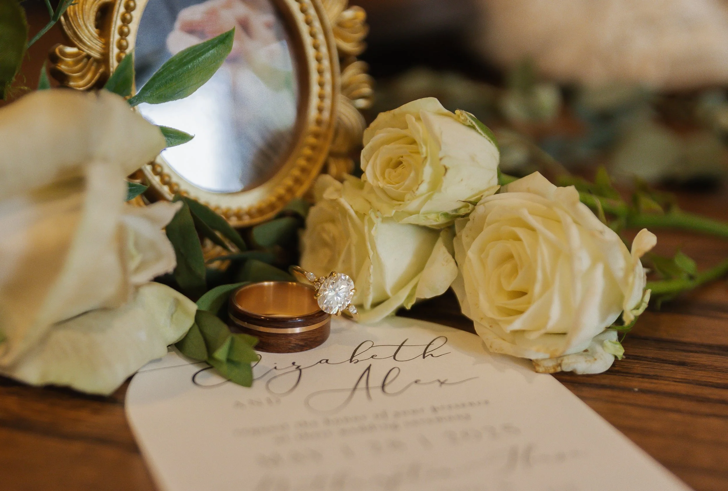 Wedding rings, white roses, a small mirror with a gold frame, and an invitation card on a wooden surface.