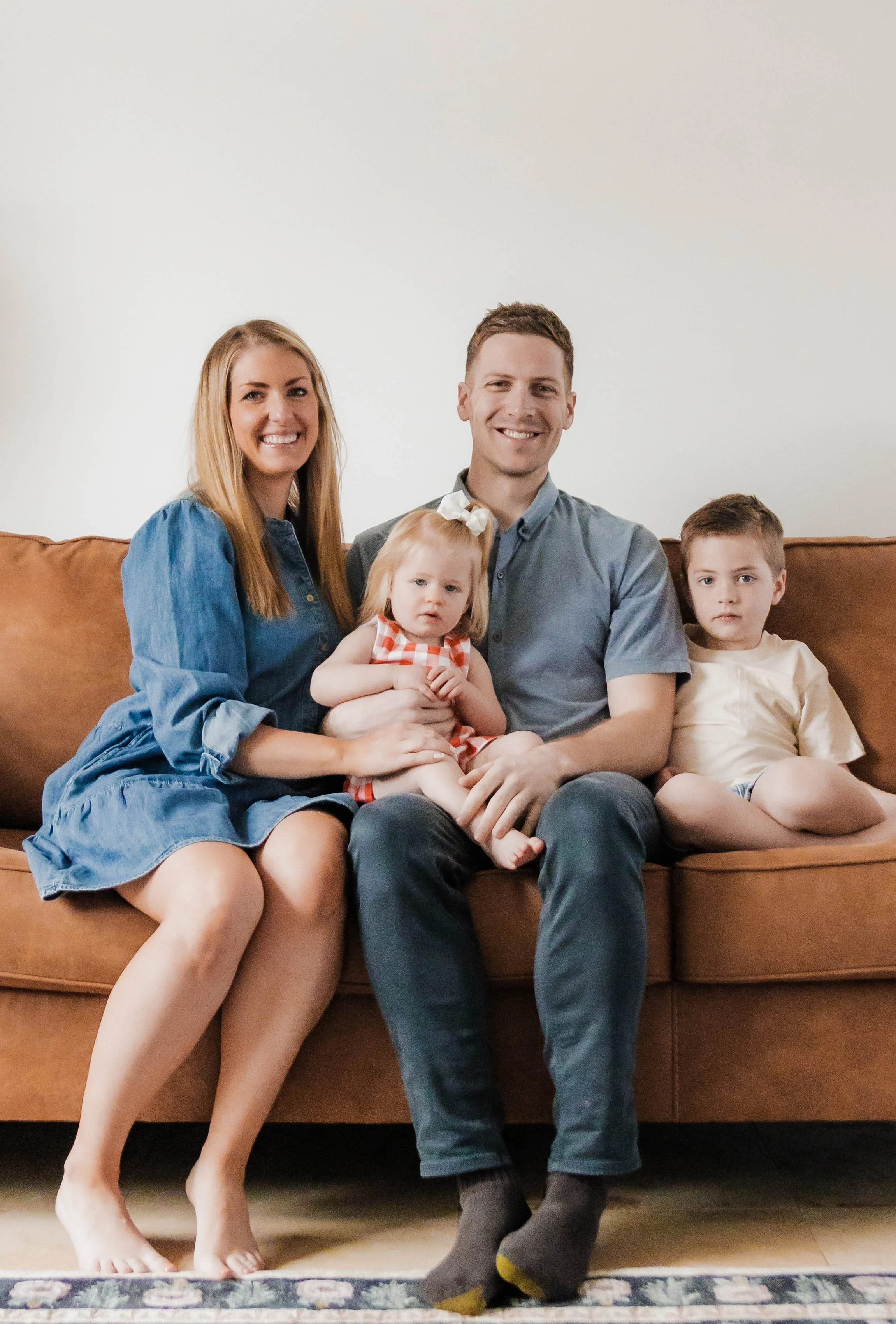 Photo of a family sitting on a leather couch, mom is blonde wearing a blue denim dress, little girl is blonde wearing a red and white dress, dad is wearing a grey shirt and jeans, little boy is wearing a white shirt and tan pants