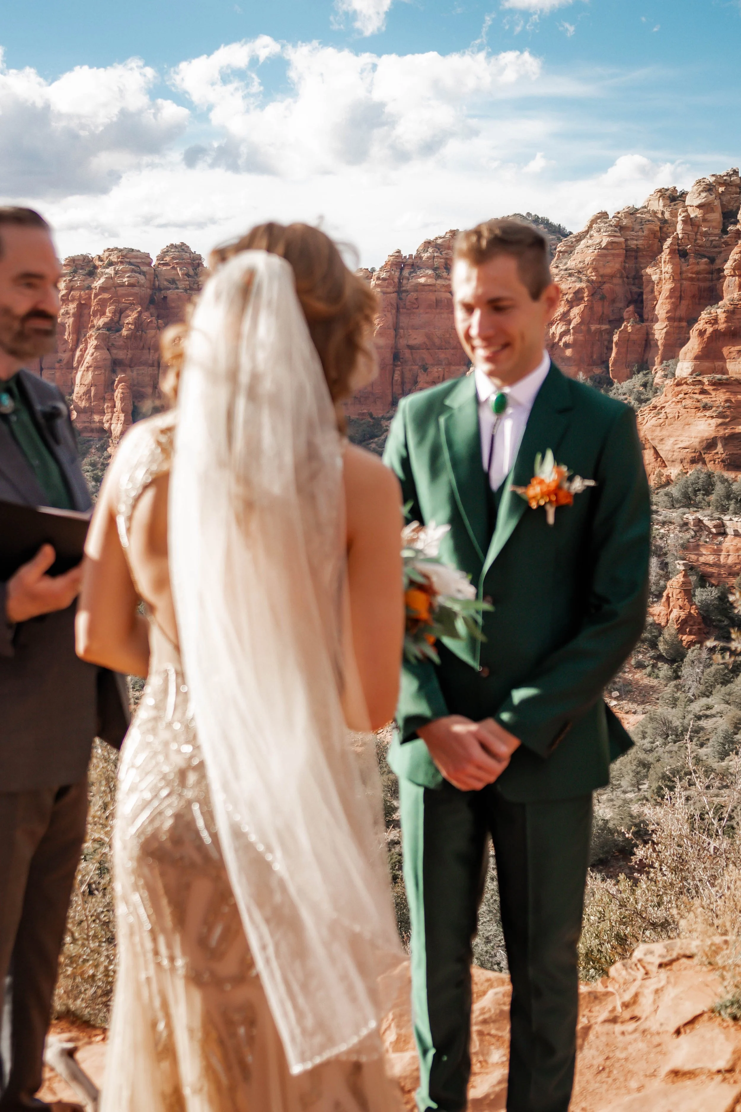 A couple getting married outdoors in front of red rock formations, with a person officiating and a scenic sky overhead.