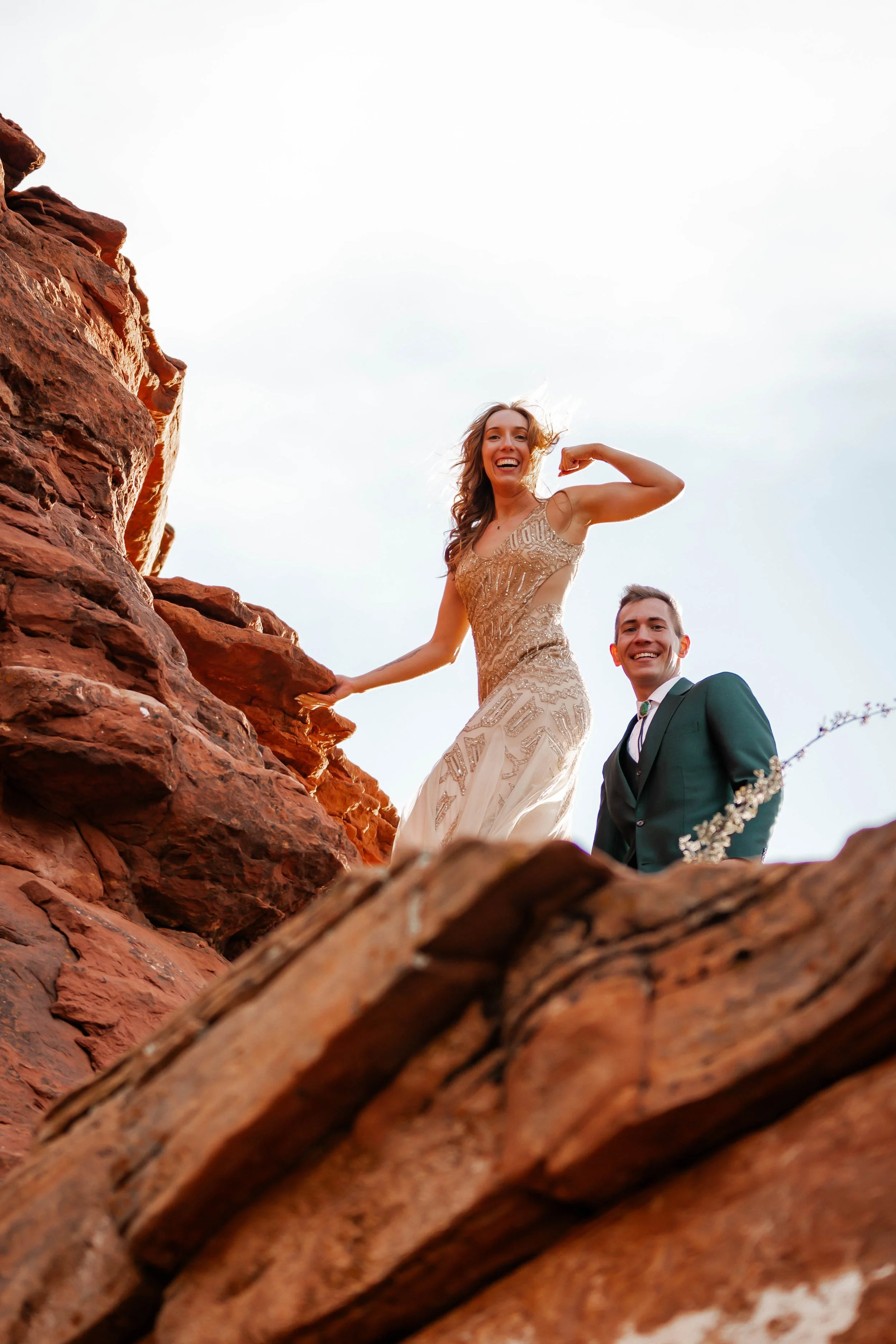 A smiling woman in an elegant dress and a smiling man in a suit posing on red rock formations outdoors.