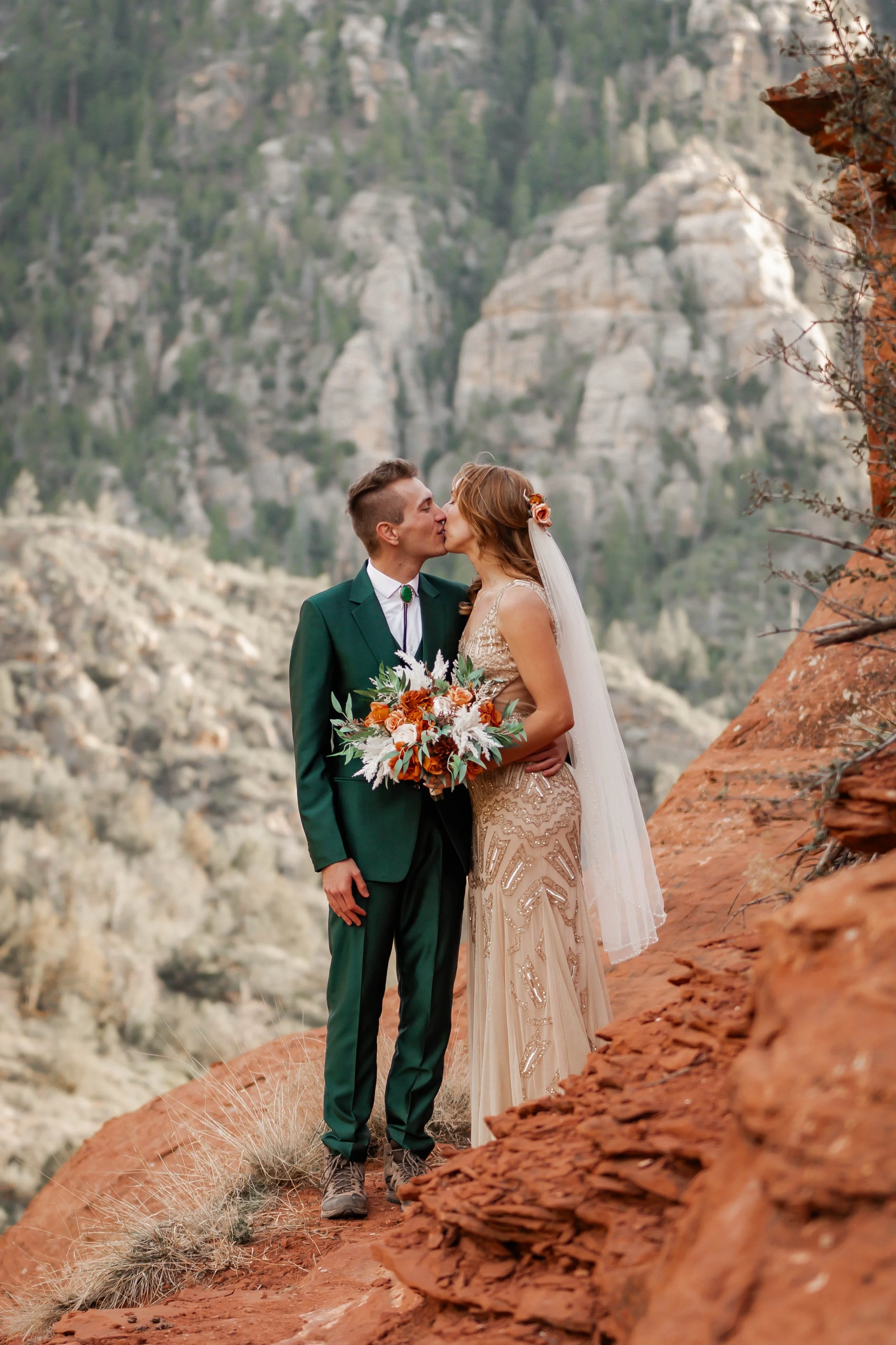A bride and groom share a kiss outdoors in a mountainous desert area, with red rocks and sparse vegetation.