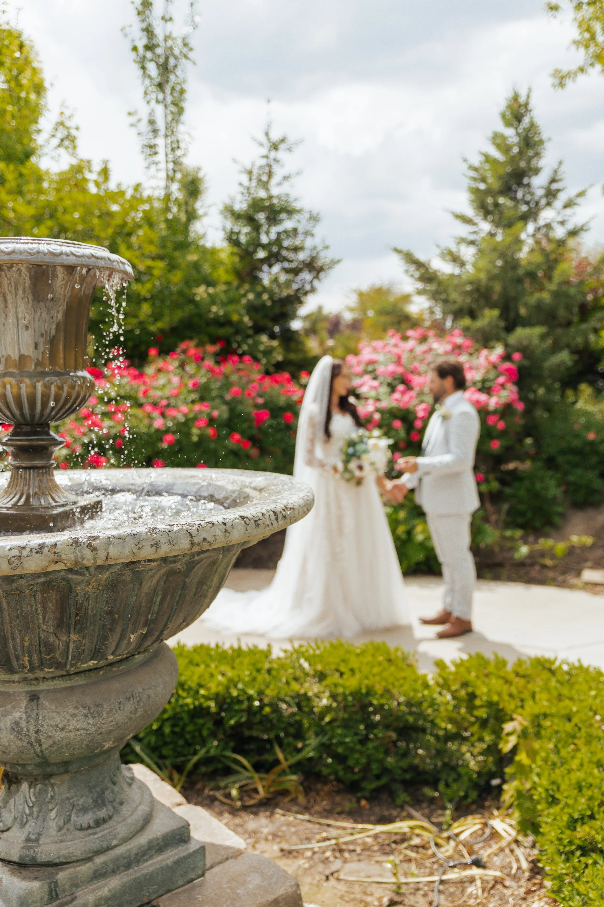 Bride and groom exchanging vows in a garden, with pink roses and green trees in background, and a fountain in the foreground.