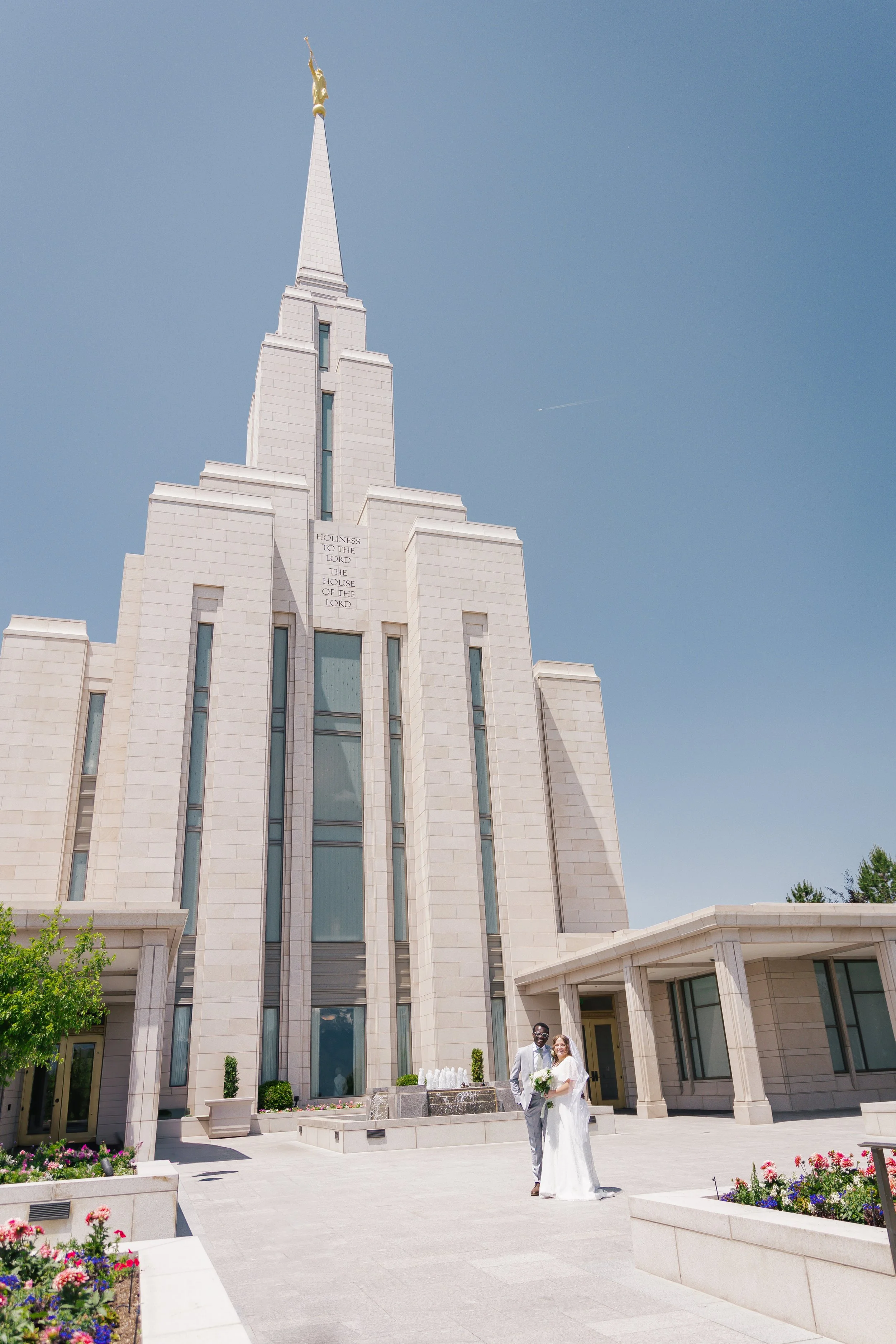 A newly married couple stands outside the oquirrh mountain temple with tall spires and a golden statue at the top, under a clear blue sky.