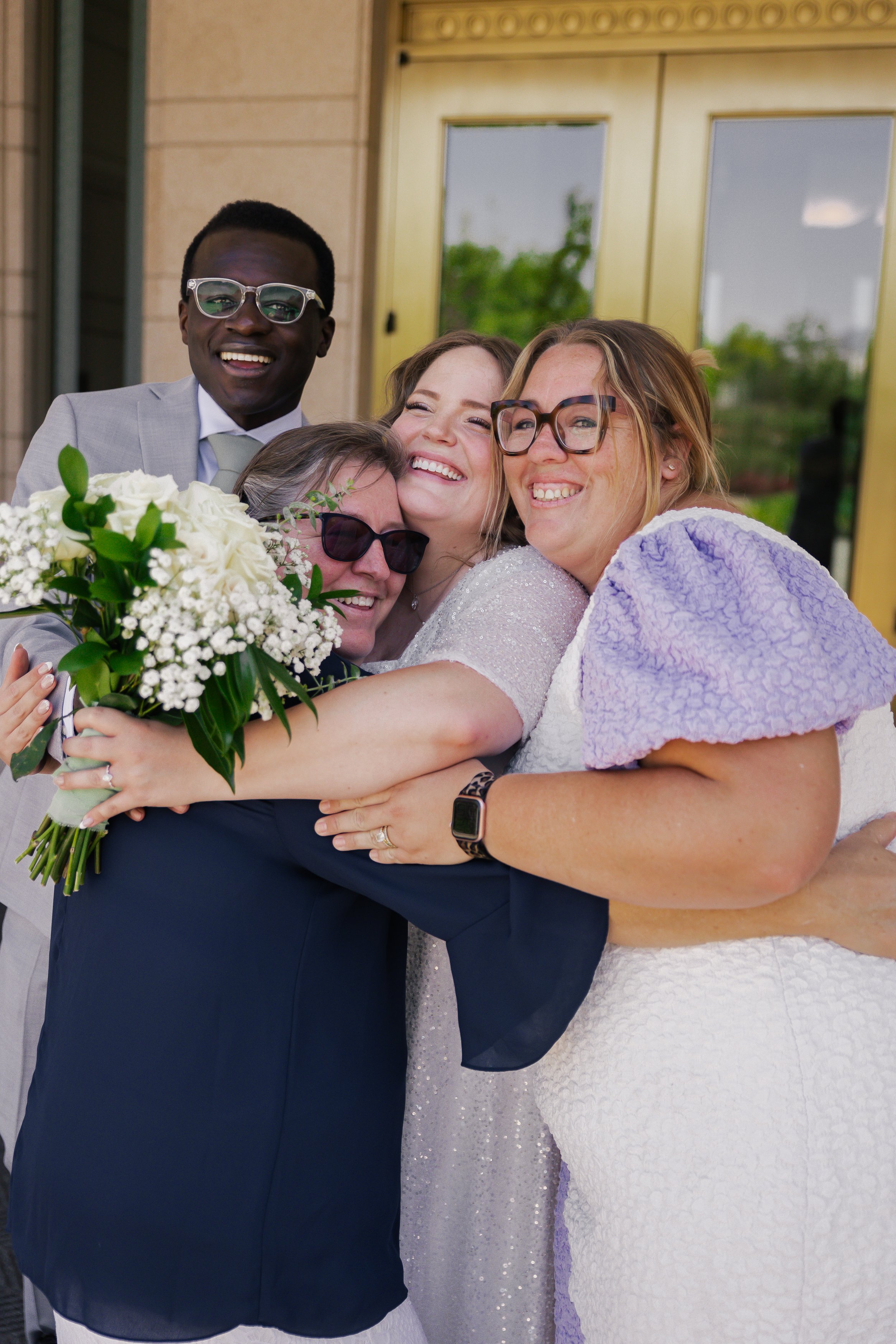 A group of five diverse women and men hugging and smiling, celebrating a special occasion, with one holding a bouquet of white flowers, standing outdoors in front of a building with glass doors.