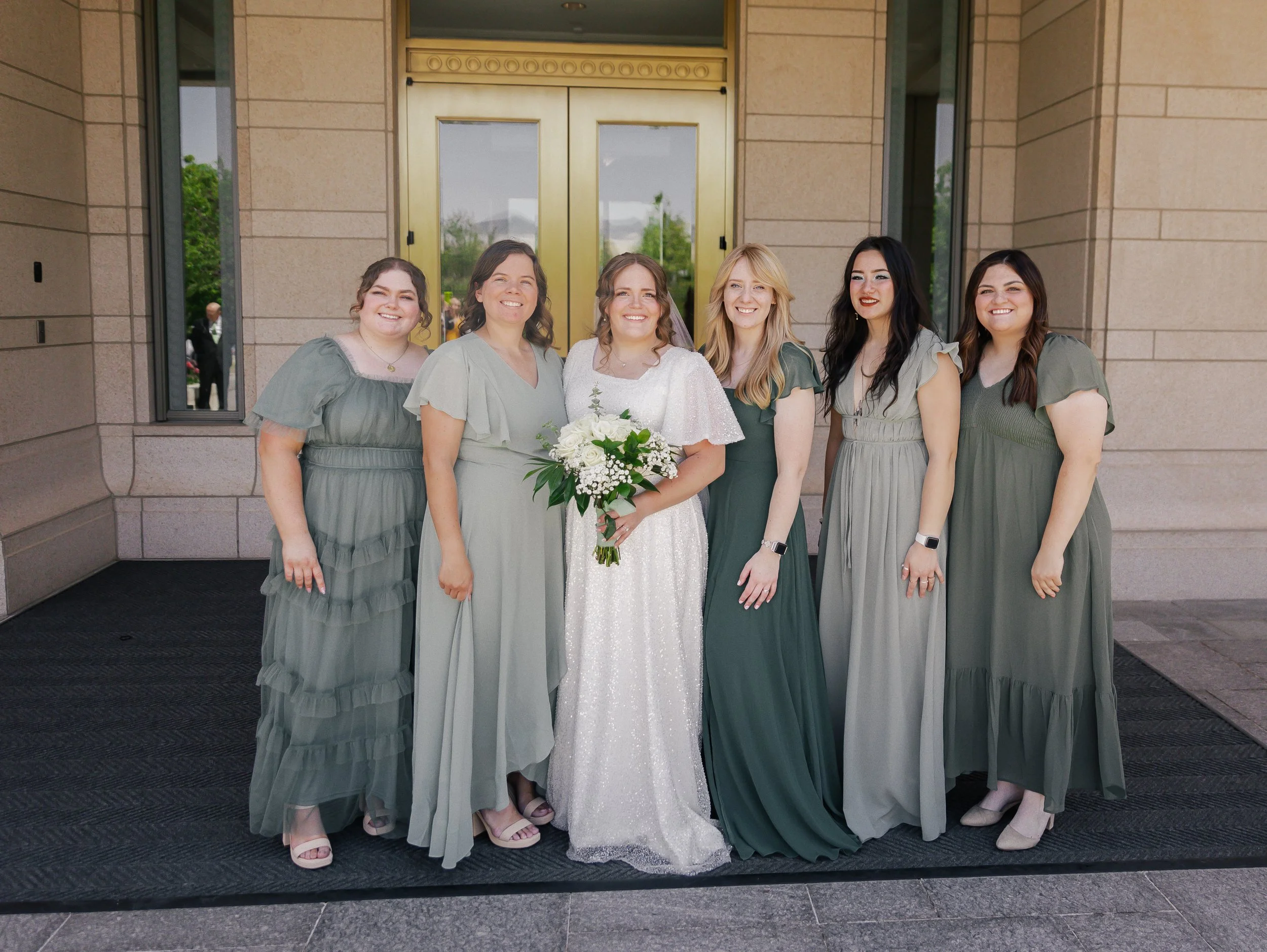 Group of seven women, including a bride in a white dress holding a bouquet, standing in front of a building entrance with gold doors, all smiling and dressed in shades of green.
