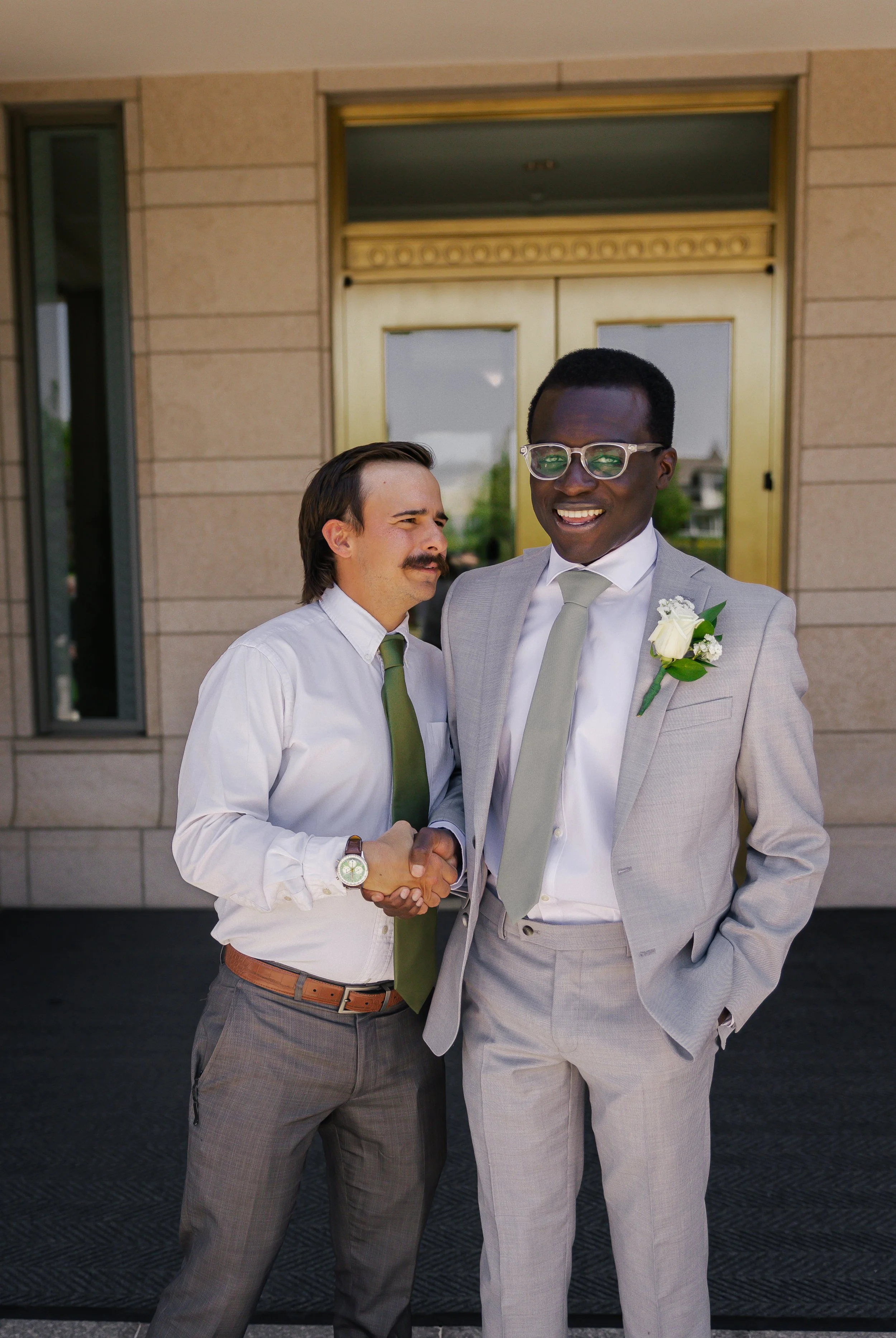 Two men in suits shaking hands and smiling outside a building.