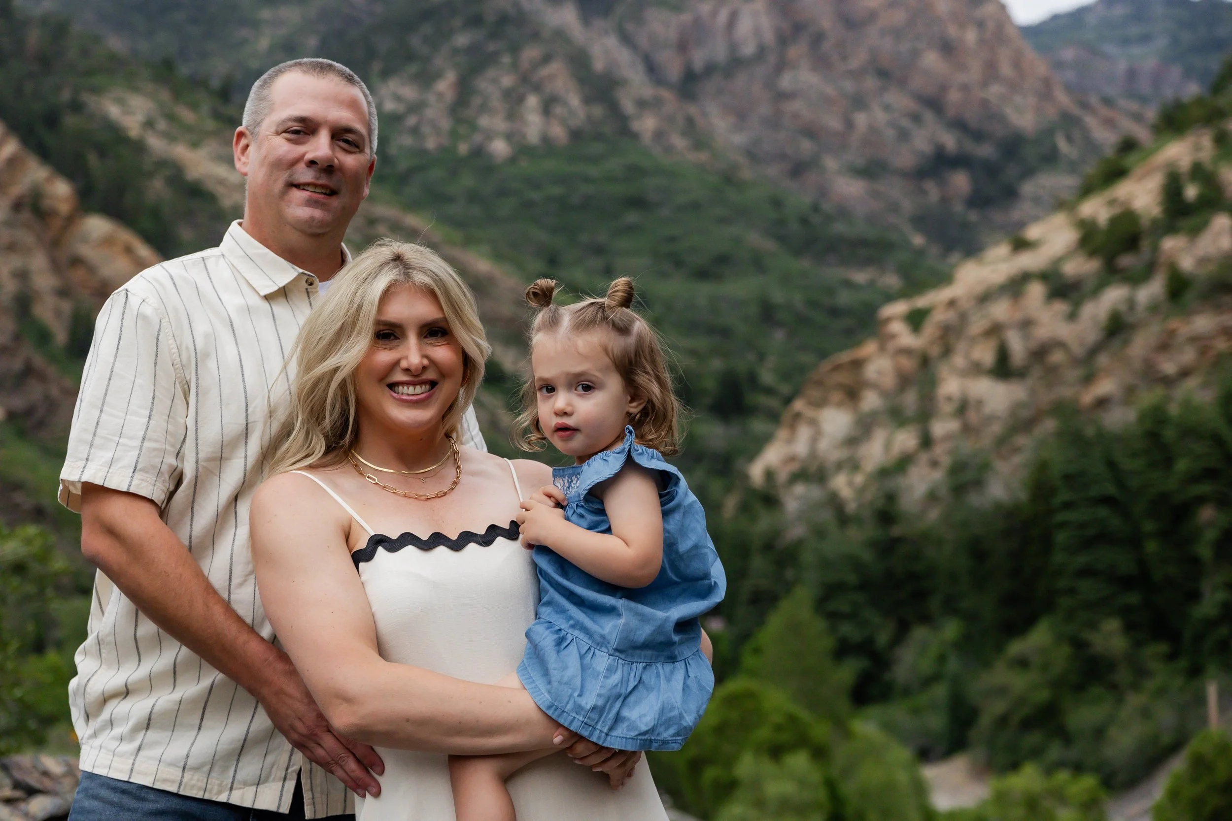 A family of three standing outdoors in a mountainous area. The woman is holding a young girl, and a man stands behind them. The background features green hills and rocky cliffs.