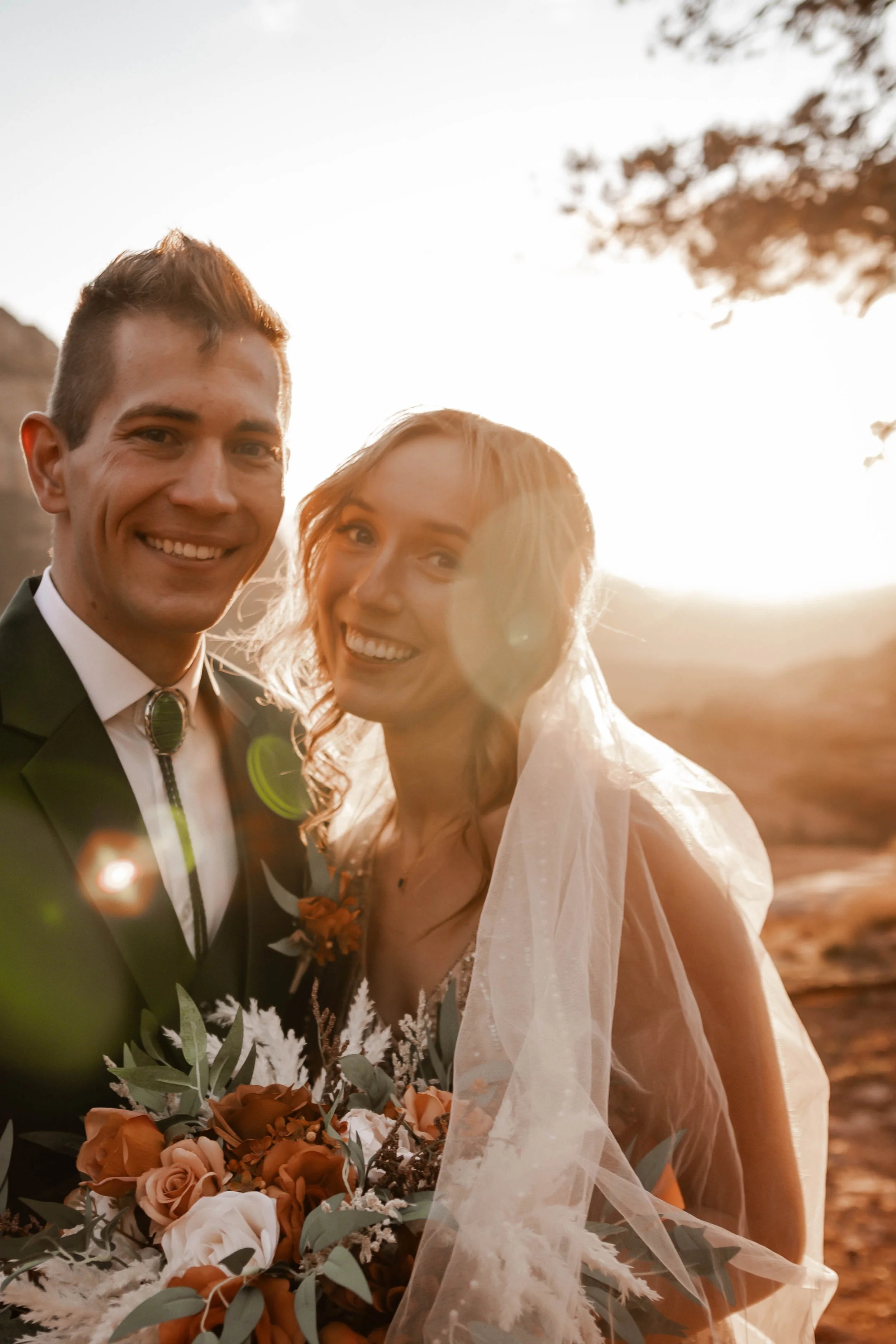 A smiling bride and groom in wedding attire outdoors at sunset, with the bride holding a bouquet of flowers and the groom wearing a black suit and bolo tie.