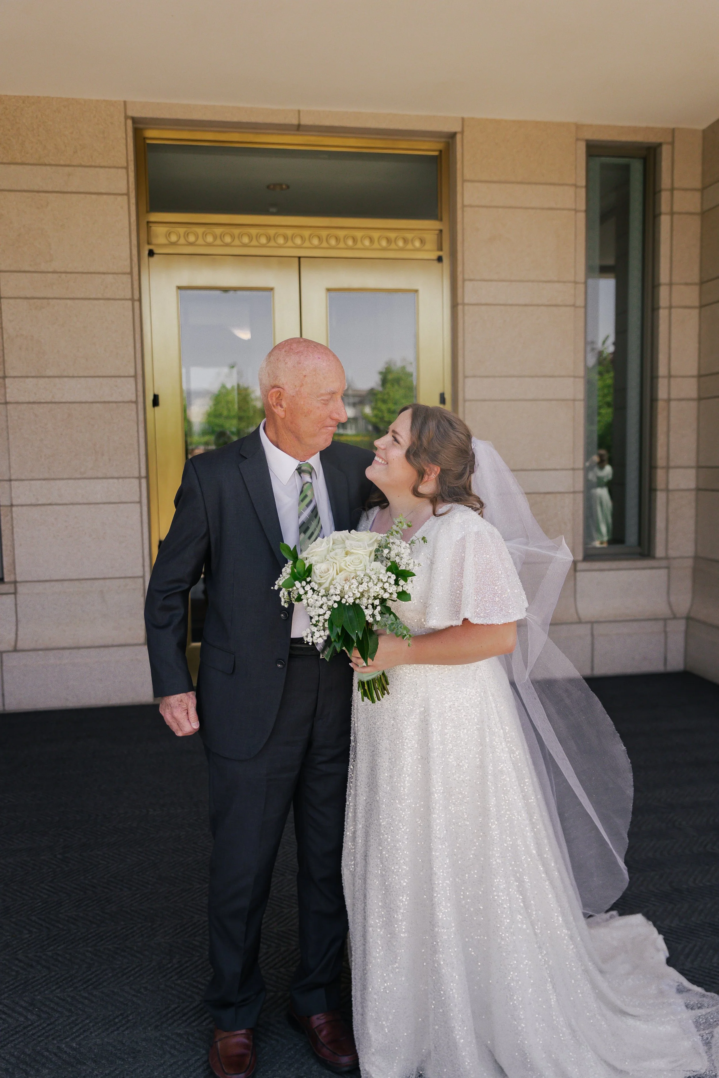 A bride and an elderly man, possibly her father, smiling and looking at each other outside a building. The bride holds a bouquet of white flowers and wears a white wedding dress with a veil. The man is dressed in a dark suit with a tie.