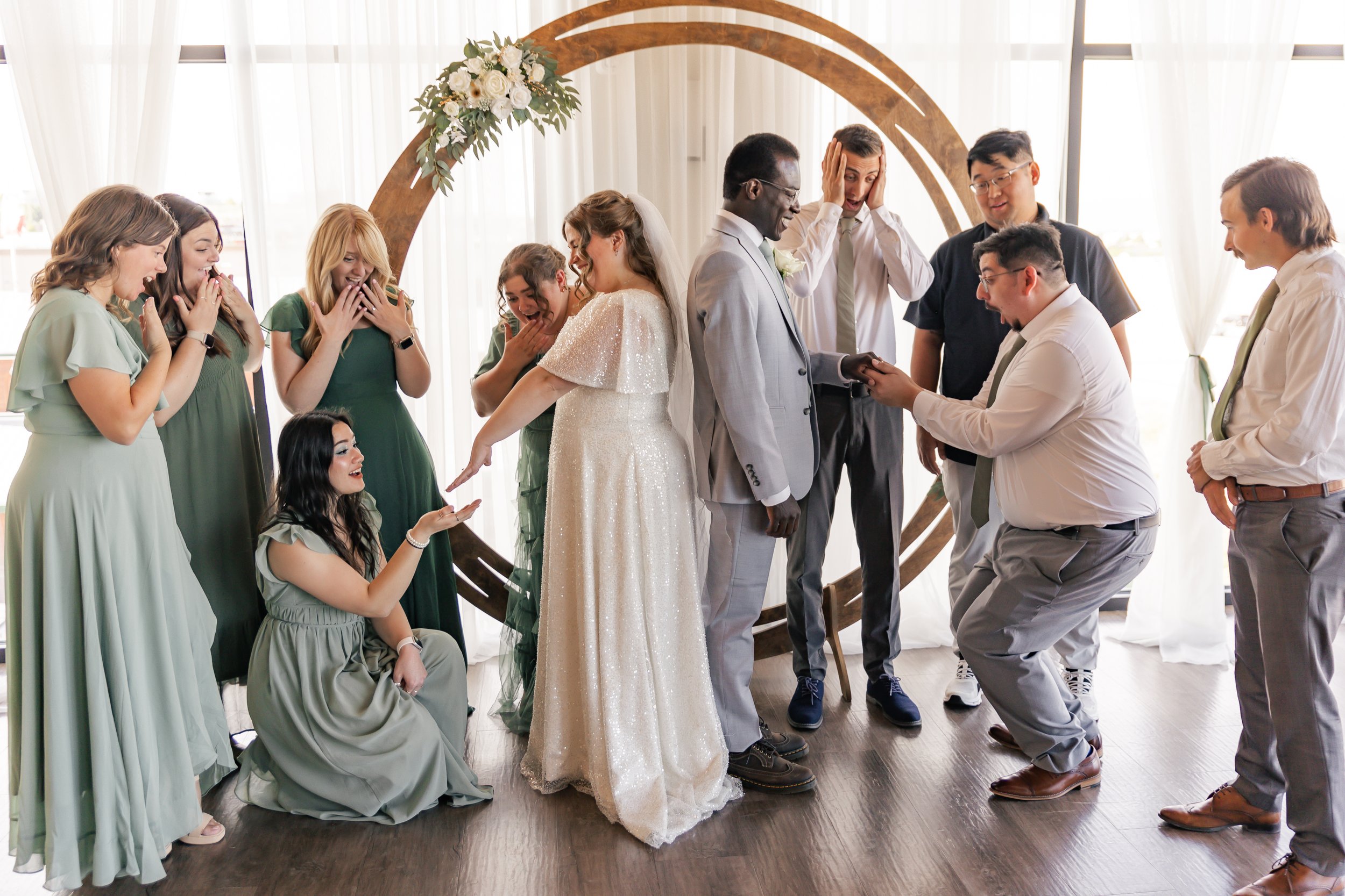 A wedding ceremony with the bride and groom exchanging rings under a wooden arch decorated with white flowers, surrounded by friends and family in a room with large windows and sheer curtains, capturing a joyful moment.