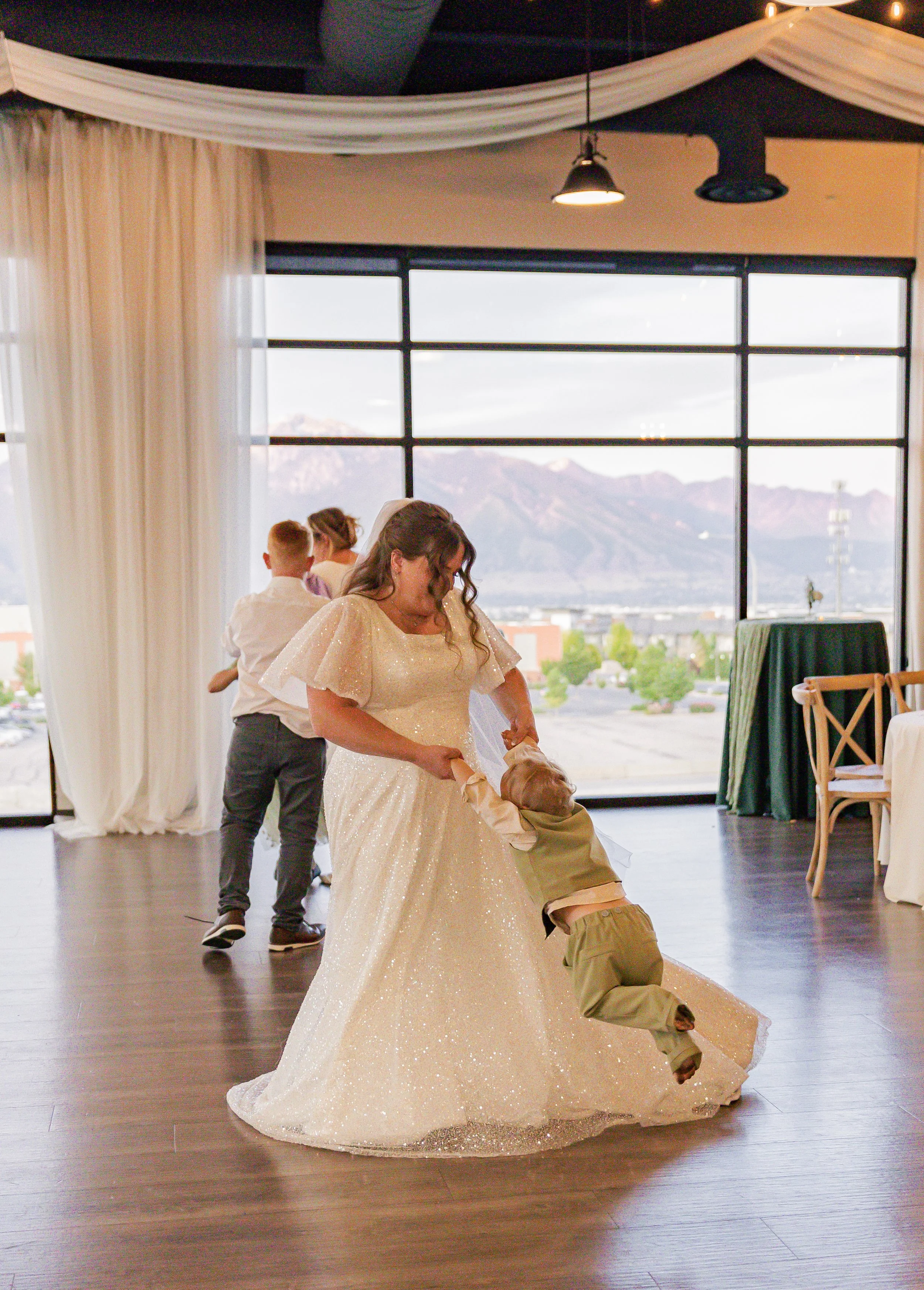 A bride dancing with a young boy at a wedding reception, with other children and large windows showing mountains in the background.