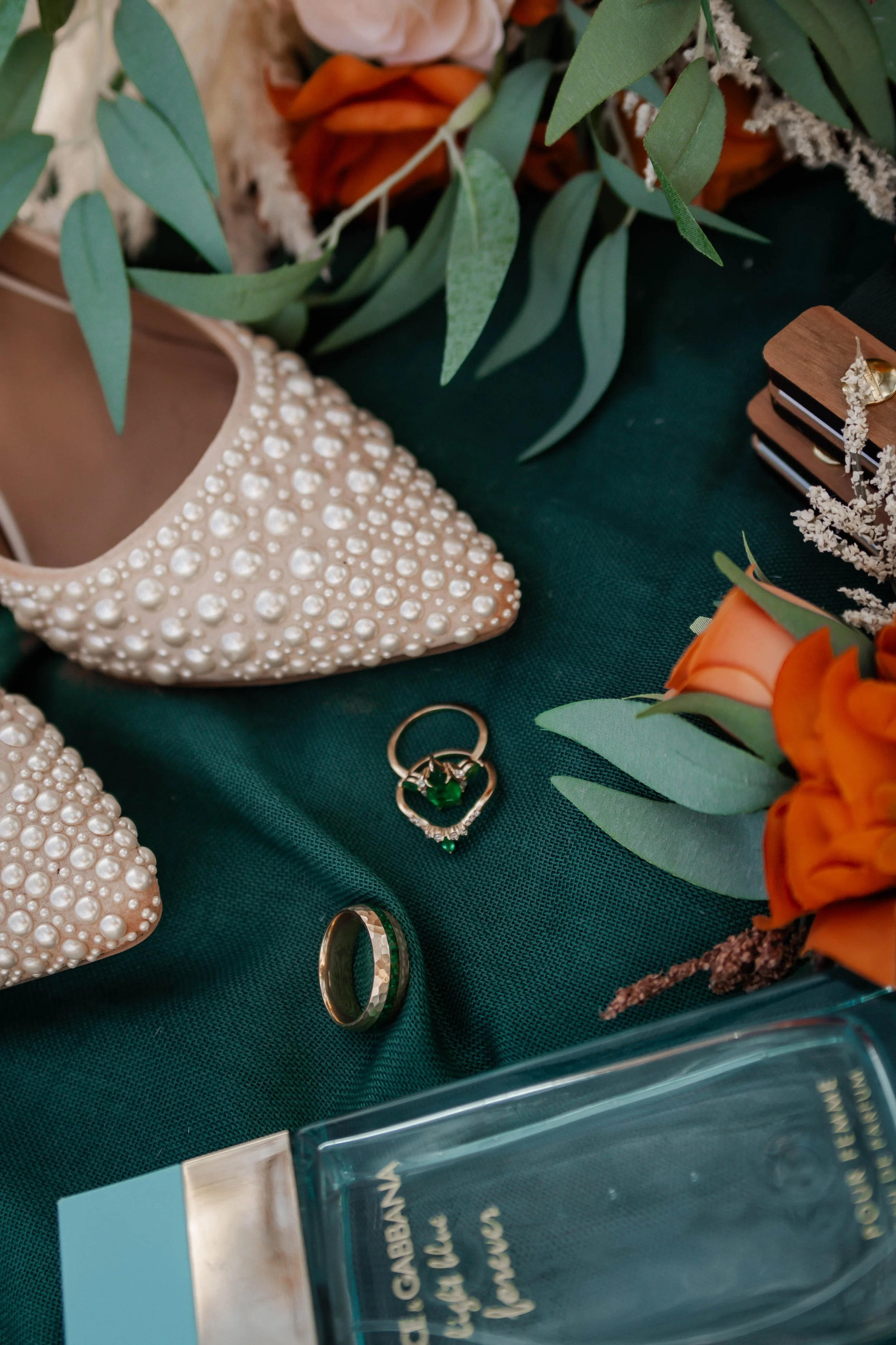 A decorative display featuring pearl-embellished beige shoes, a gold wedding ring, a ring with a green gem, a floral bouquet with orange roses and greenery, and some jewelry and perfume bottles on a dark green cloth.
