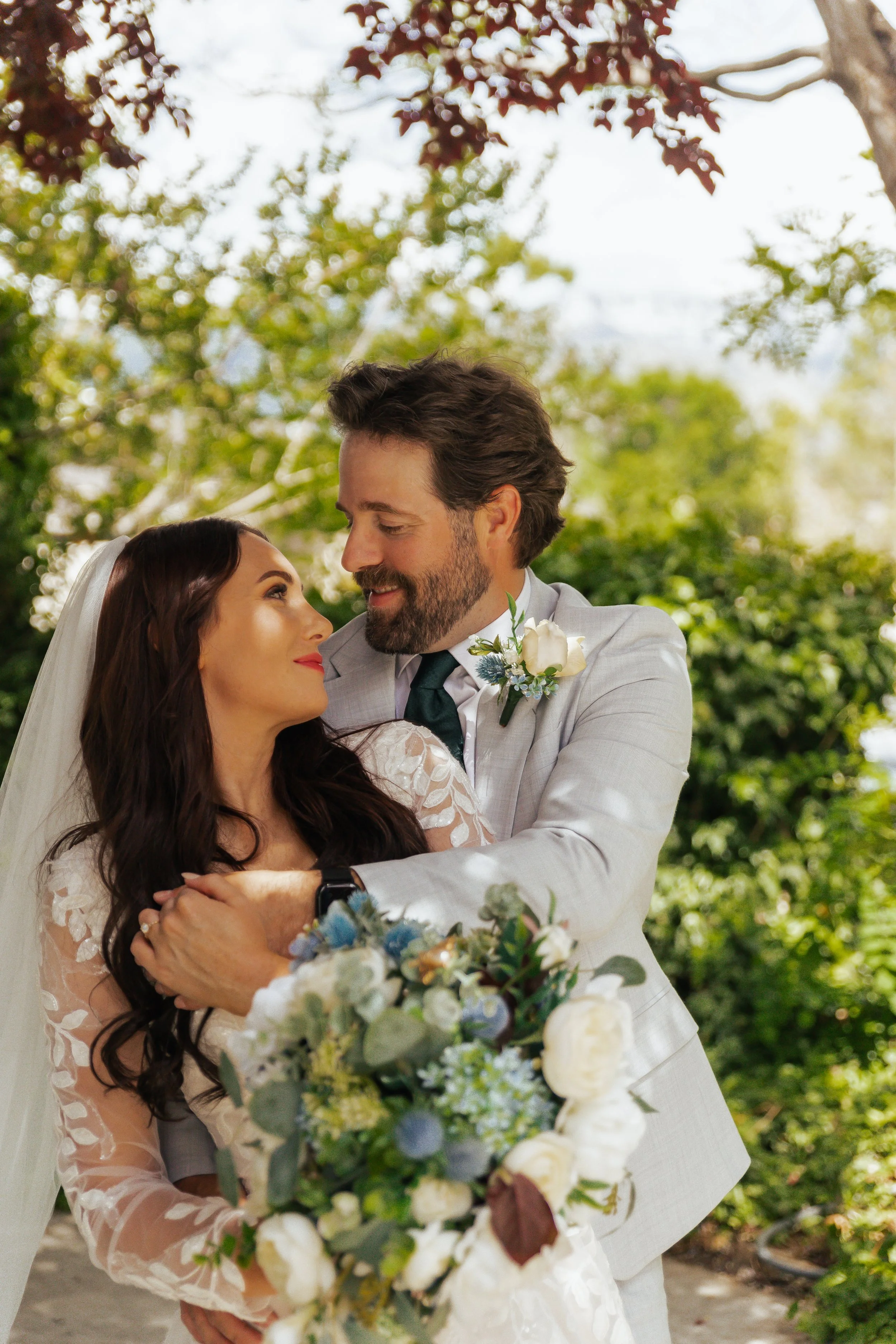 A bride and groom embracing outdoors, with green trees in the background, during their wedding.