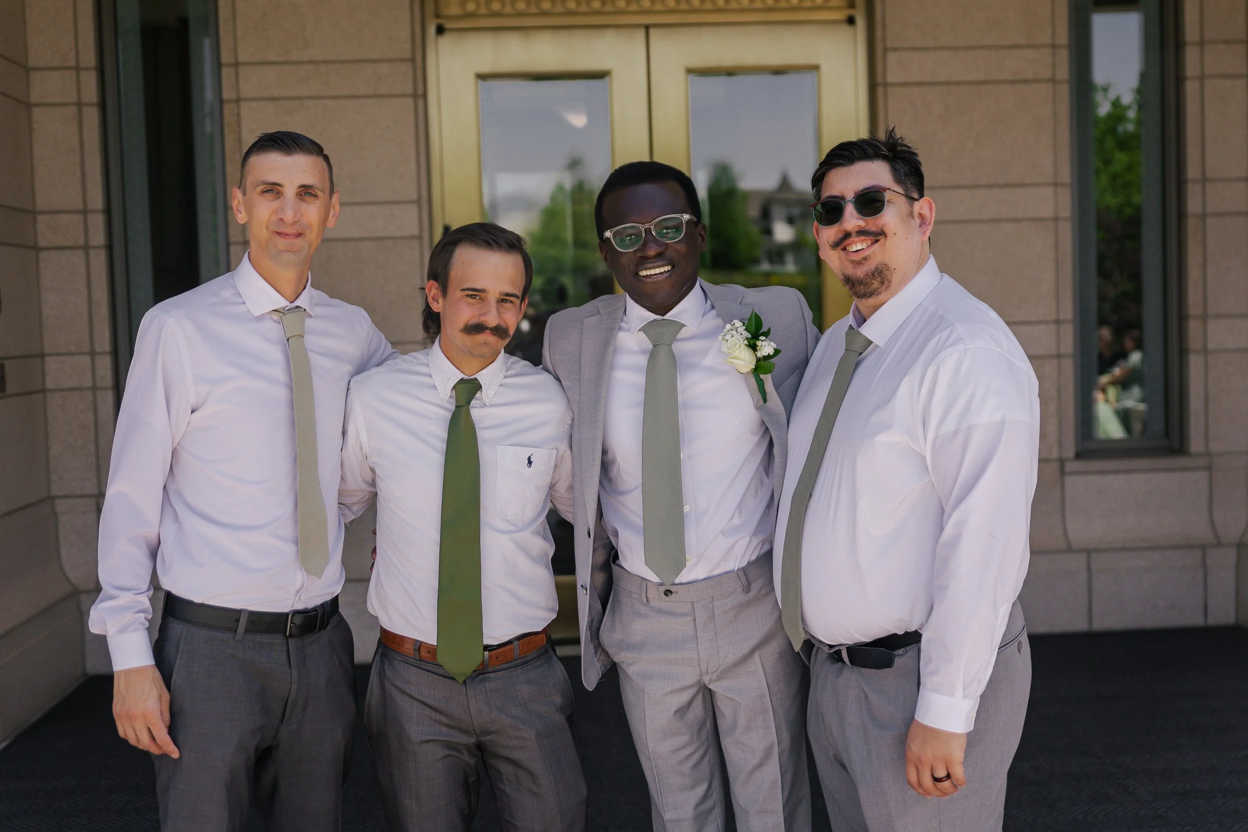 A group of five men in formal attire standing outside a building, smiling for the photo. They are dressed in white shirts with gray ties and gray pants, with one man in a light gray suit jacket. The man in the center is wearing sunglasses and a bouto