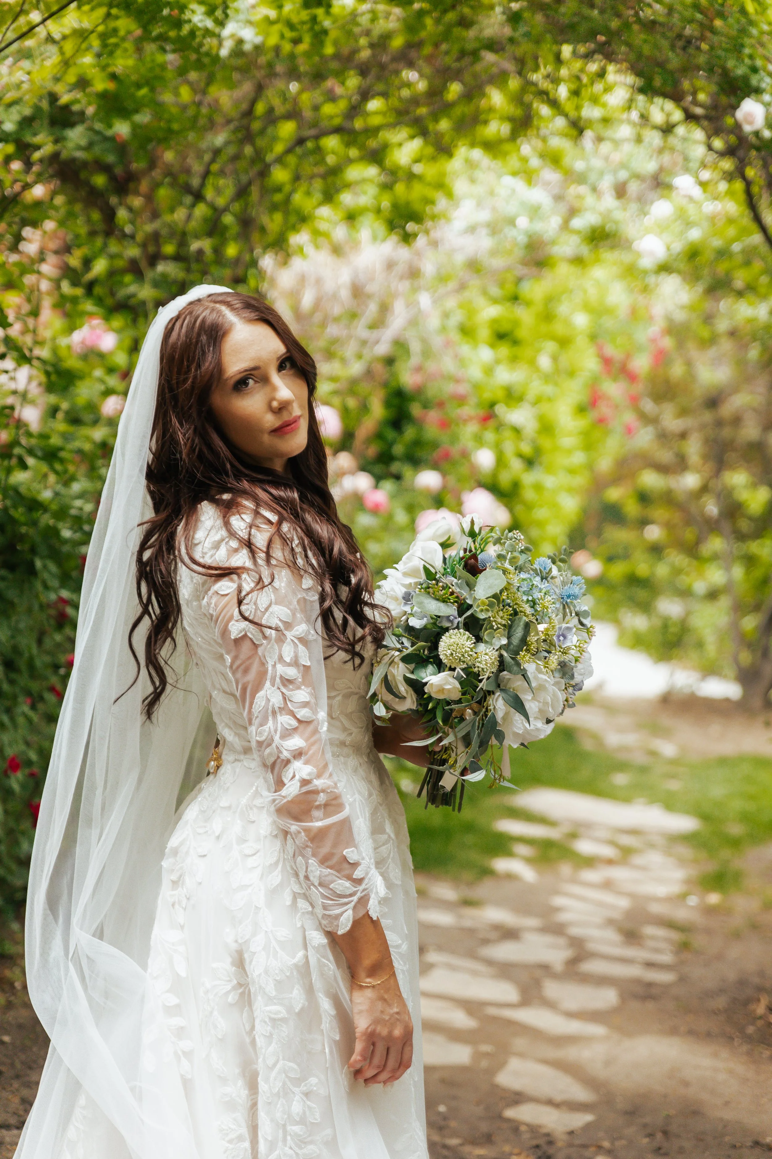 A bride holding a bouquet of white and blue flowers standing on a garden path surrounded by green trees and blooming flowers.