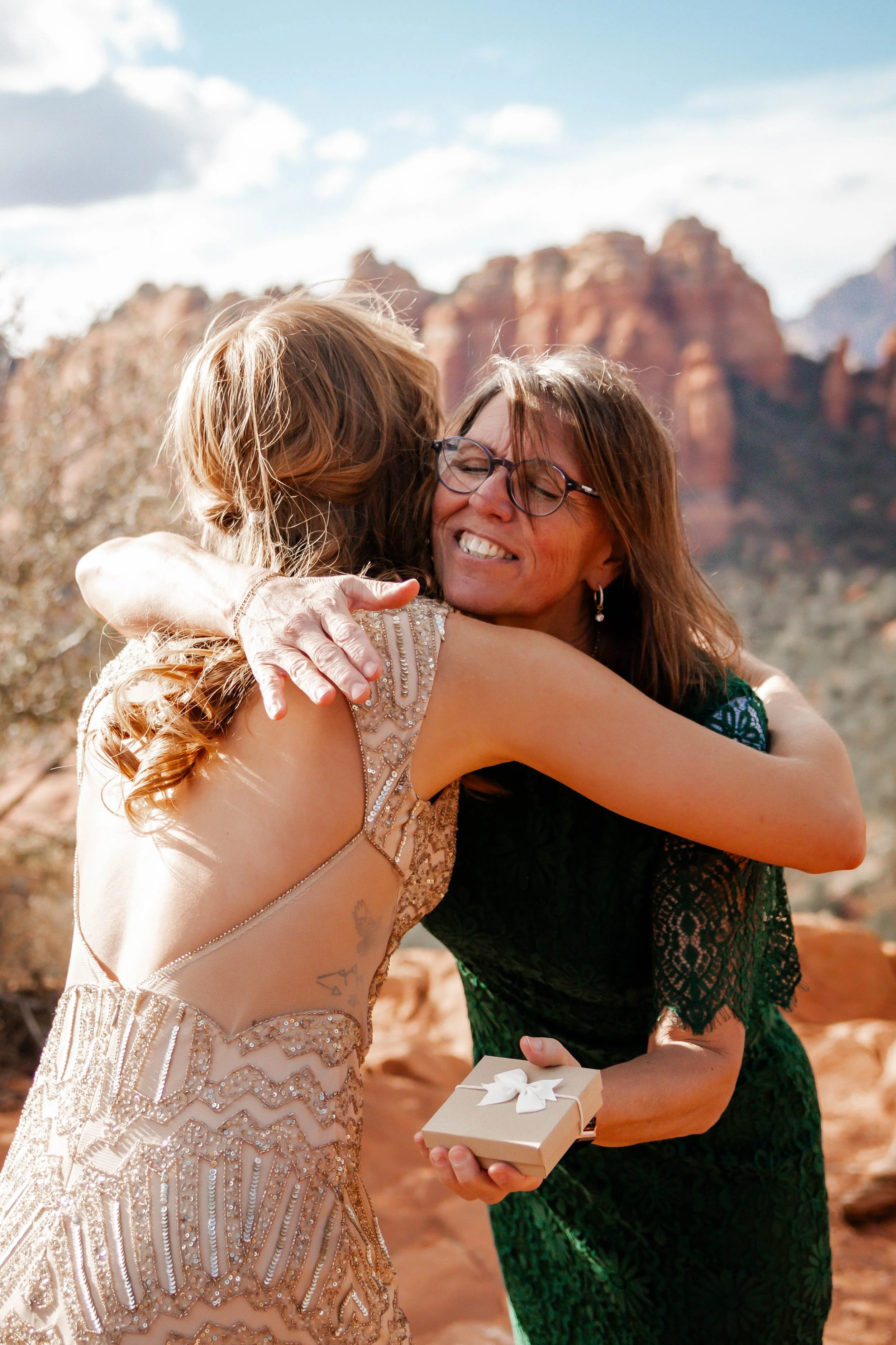 Two women hugging outdoors, one in a light-colored, embellished dress and the other in a dark green lace dress, with a mountain landscape in the background.