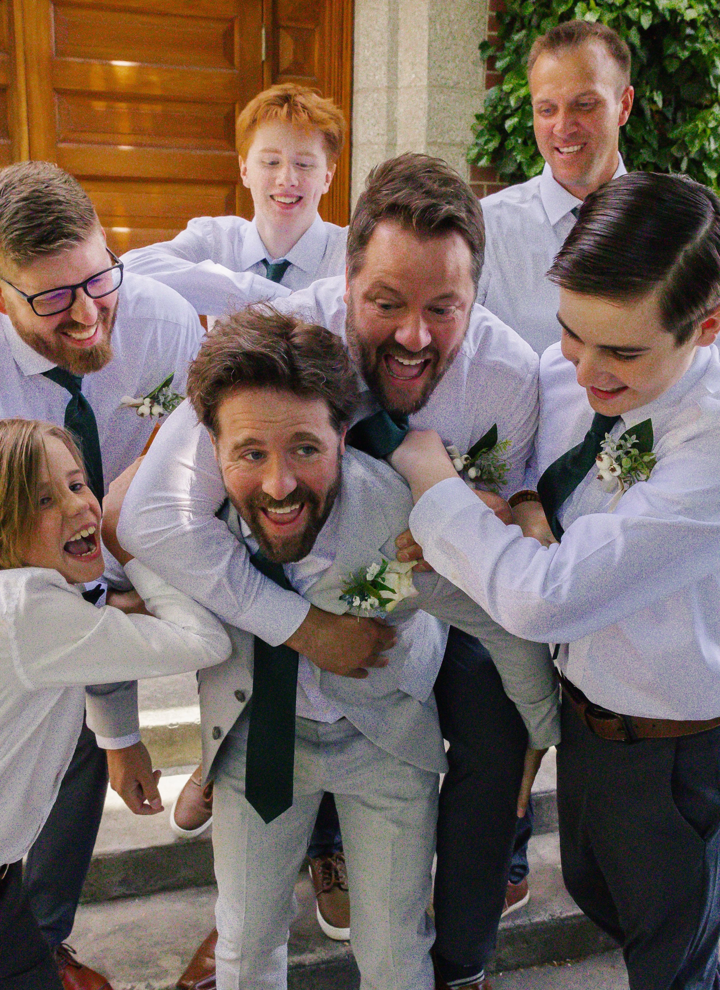 Group of people, mostly men, celebrate and hug each other outside a building on a wedding day, wearing formal attire with flowers on their shirts and ties.