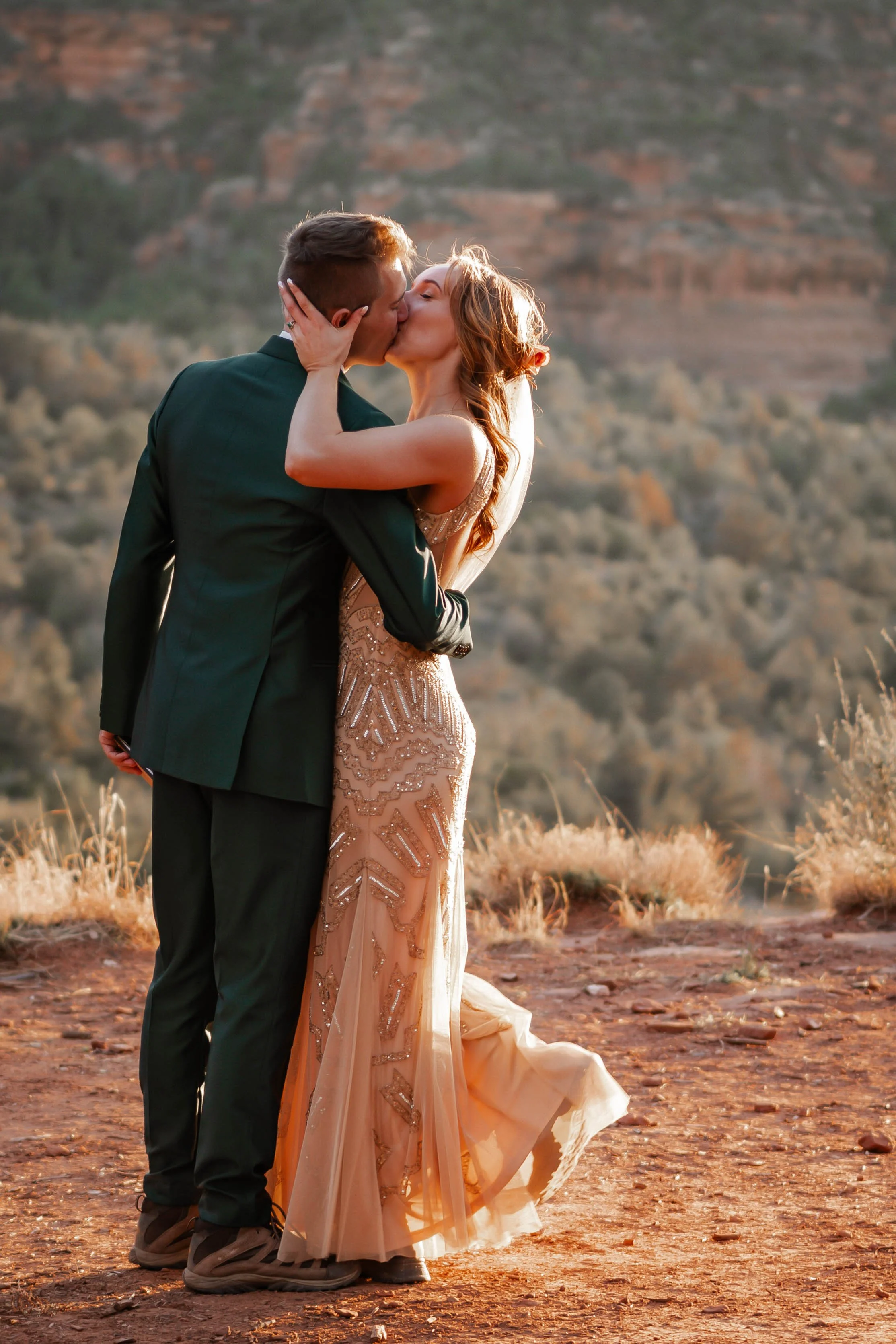 A couple in wedding attire sharing a kiss outdoors at sunset with a mountainous desert background.