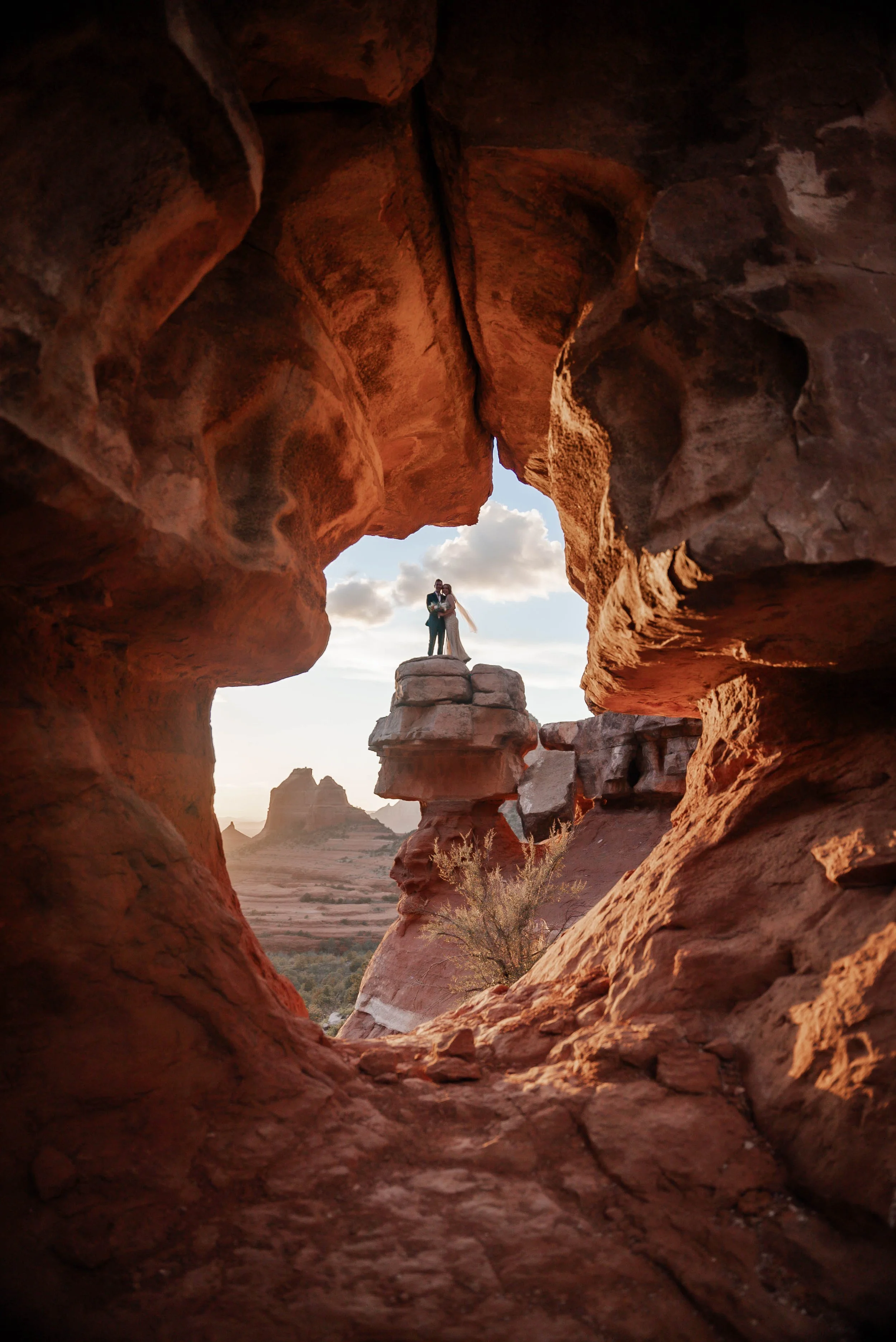 A couple dressed in wedding attire standing together on a rock formation in a desert landscape, seen through a natural rock window known as merry go round rock in Sedona, Arizona.