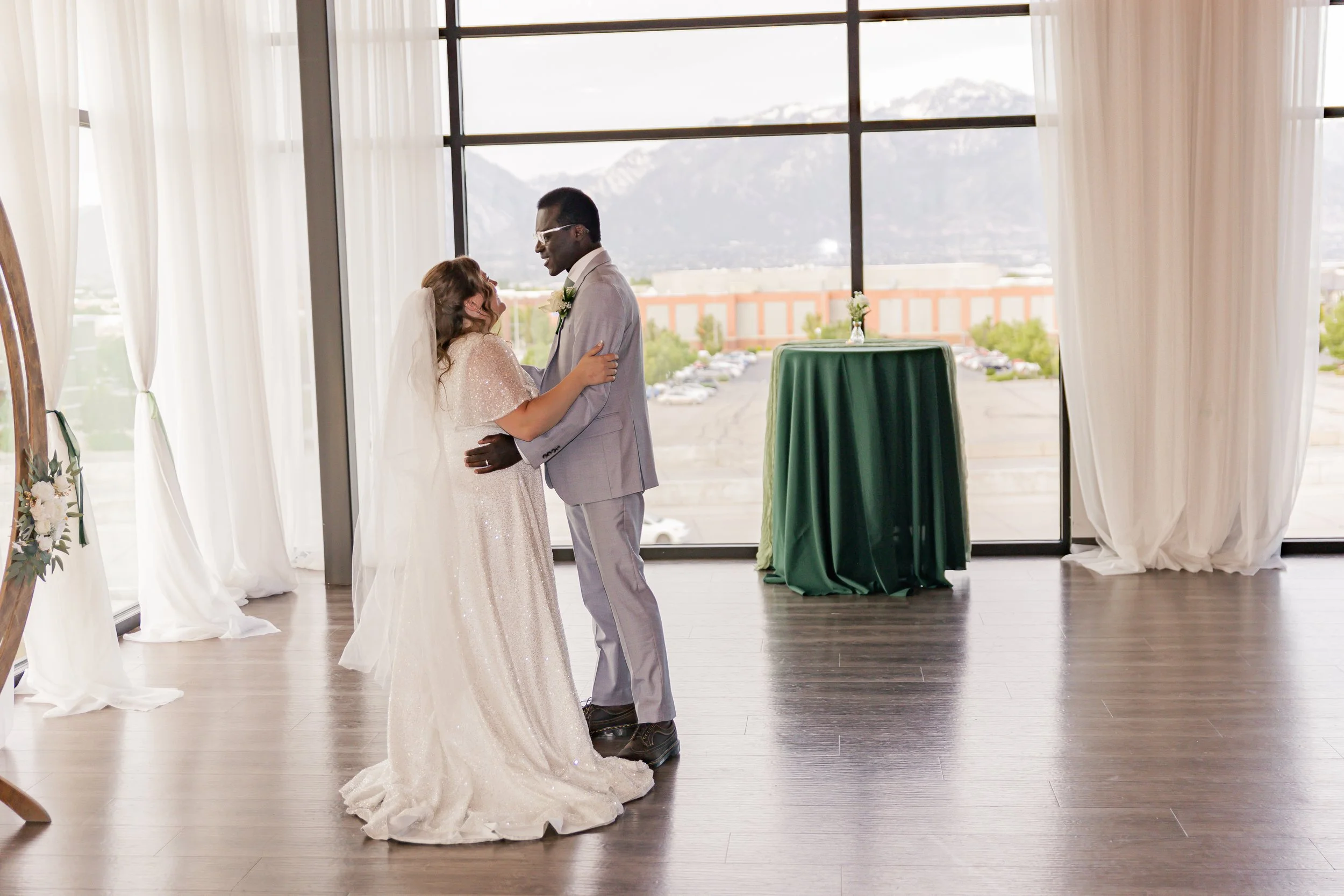 A bride and groom dancing during their wedding ceremony indoors near large windows with mountain scenery in the background.