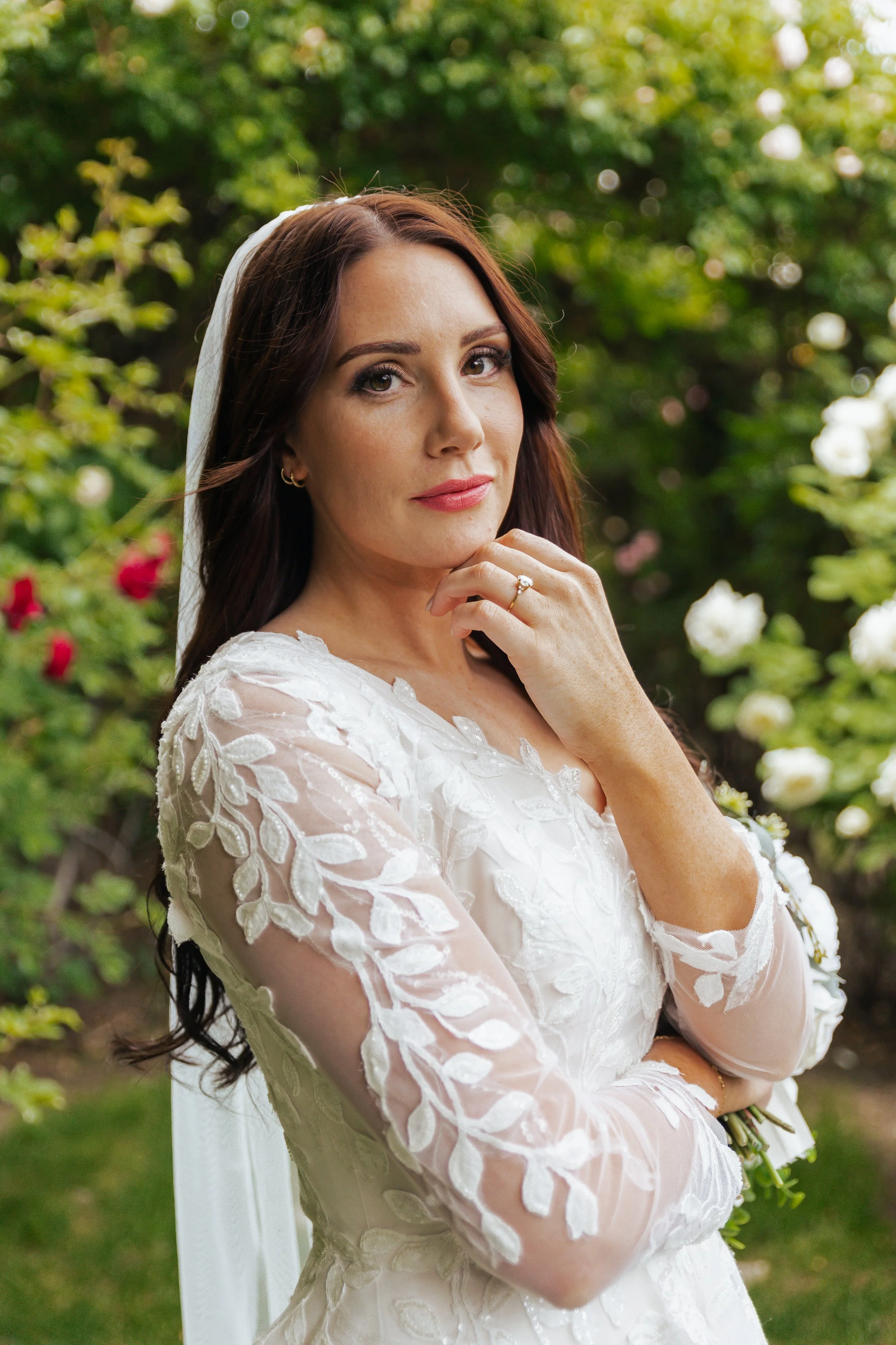 Portrait of a bride in a white lace dress outdoors surrounded by greenery and flowers.