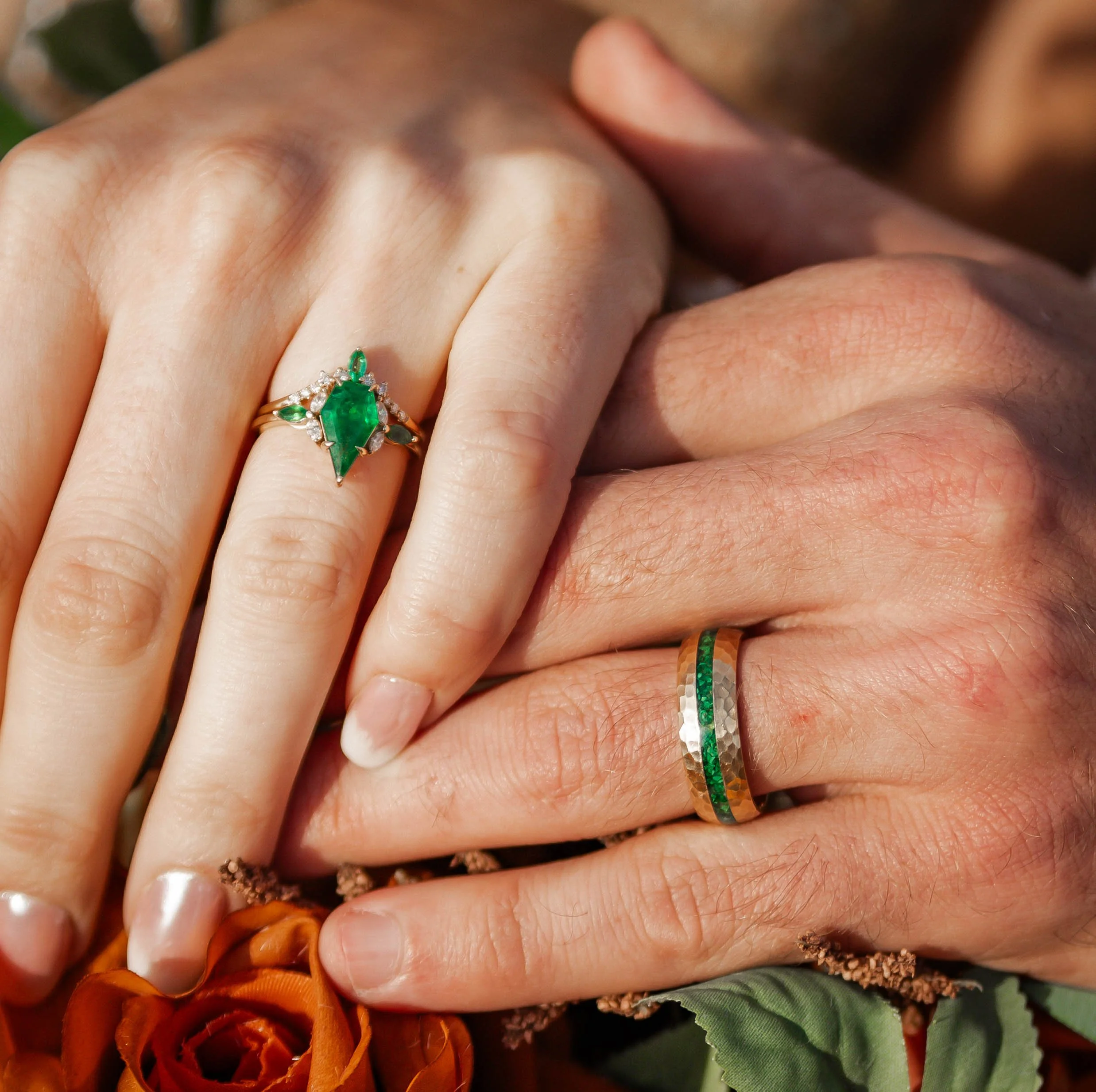 Close-up of two hands with wedding rings, one with a green gemstone and diamonds, the other with green, silver, and gold bands, resting on a bouquet of orange flowers.