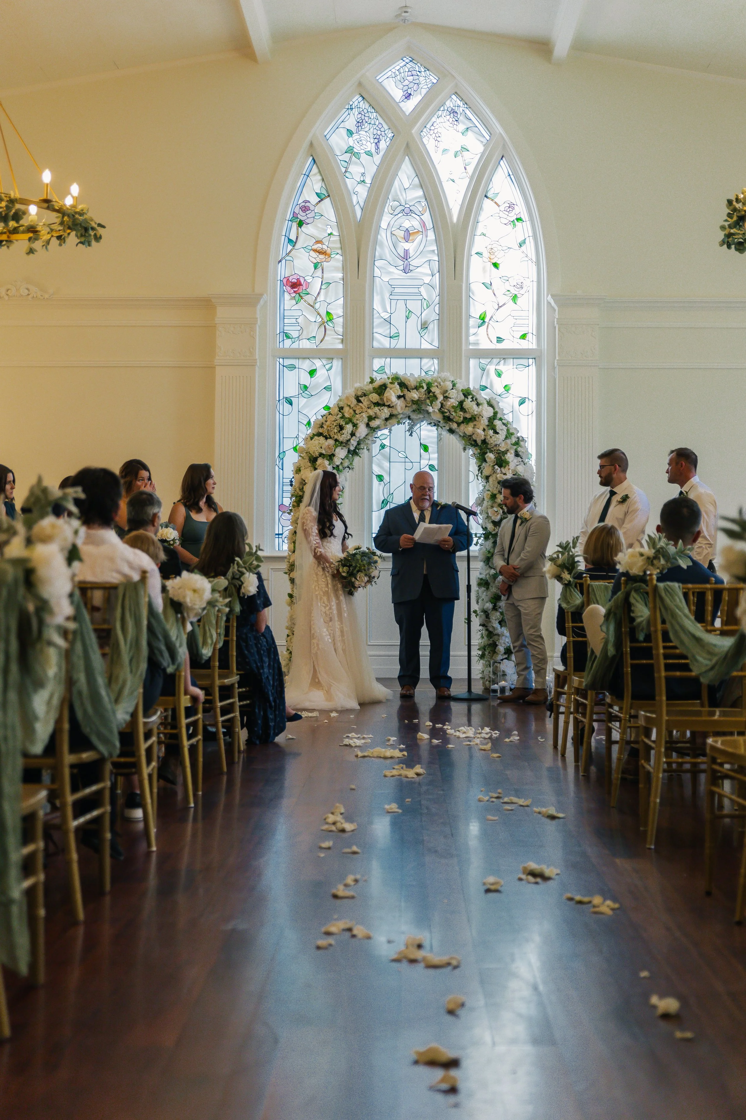 A wedding ceremony with a bride and groom standing under a floral arch in front of an officiant, inside a church with stained glass windows. Guests sit on both sides of the aisle decorated with flowers and draped fabric.