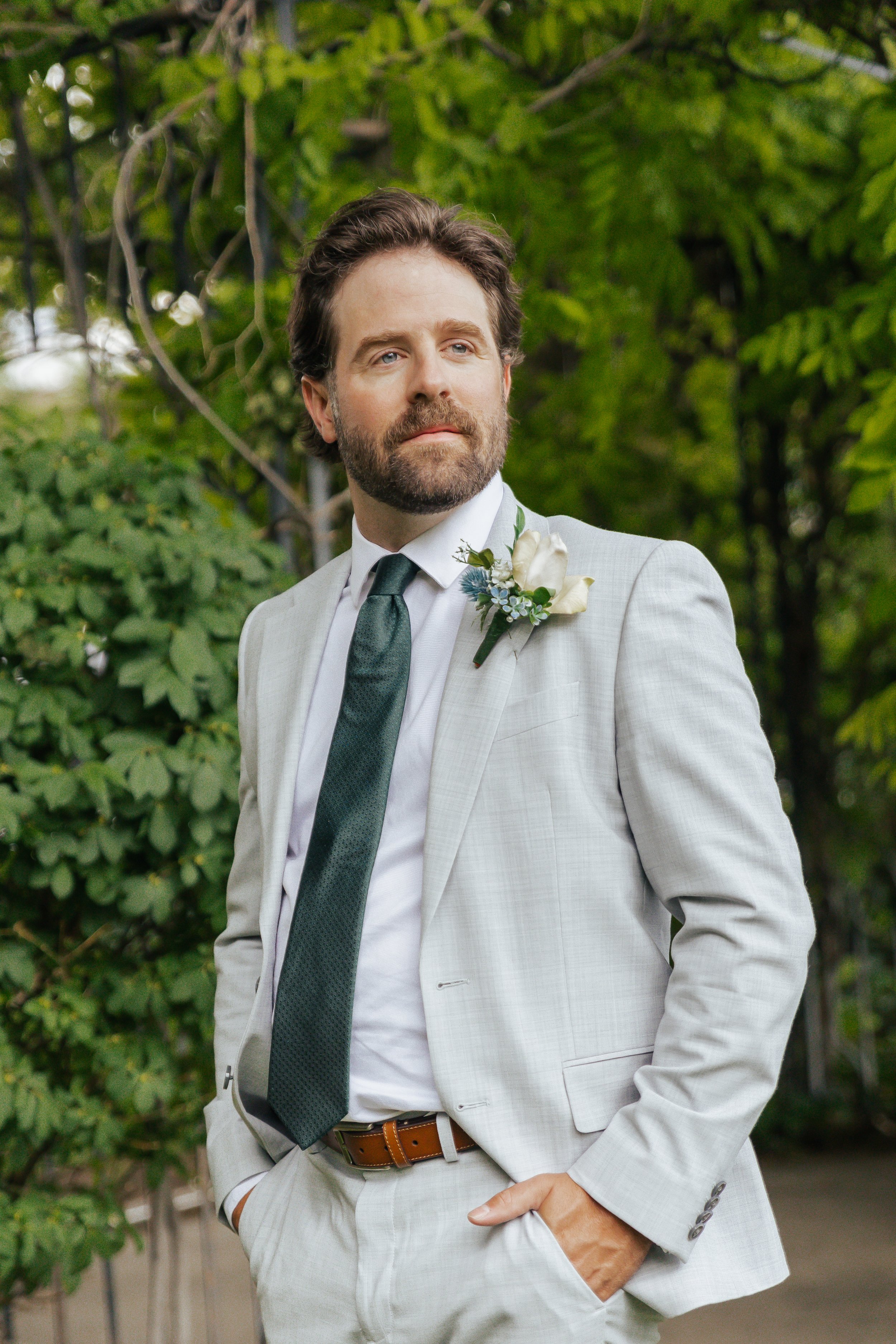 A man in a light gray wedding suit with a dark green tie and boutonniere, standing outdoors with trees in the background.