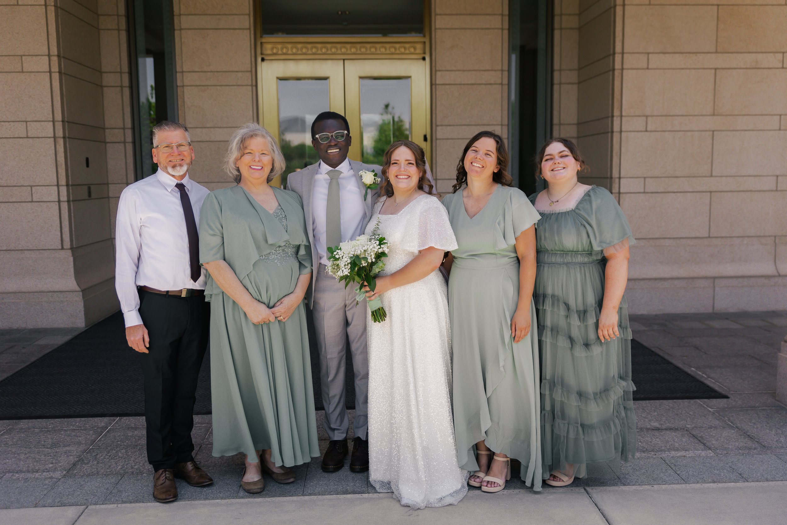 Group of six people, including a bride holding a bouquet, standing outside a building with stone walls, smiling for a wedding photo.