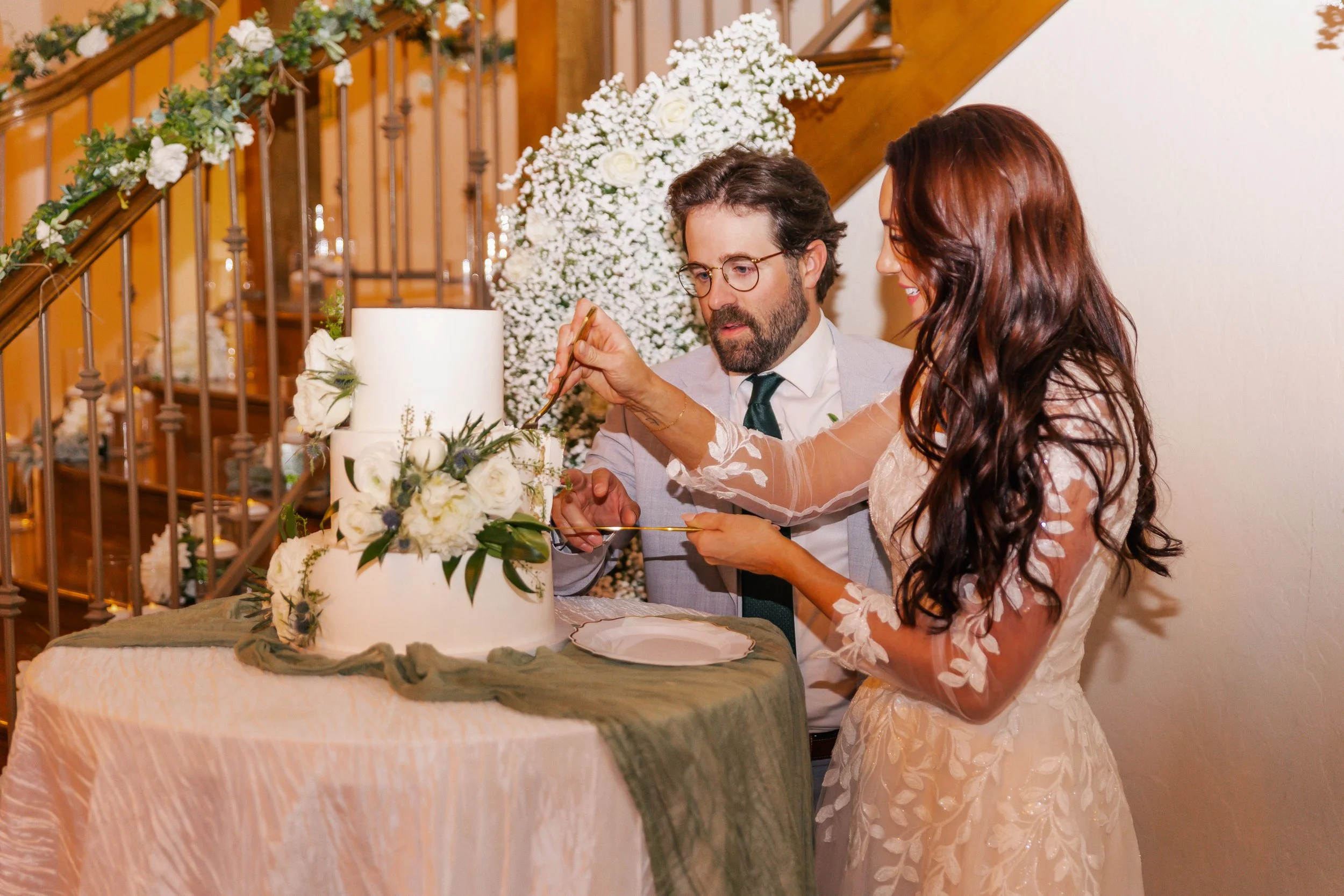 A bride and a man, possibly the groom, are cutting a wedding cake decorated with white flowers and greenery. The scene is set at a wedding reception with floral arrangements and a staircase in the background.