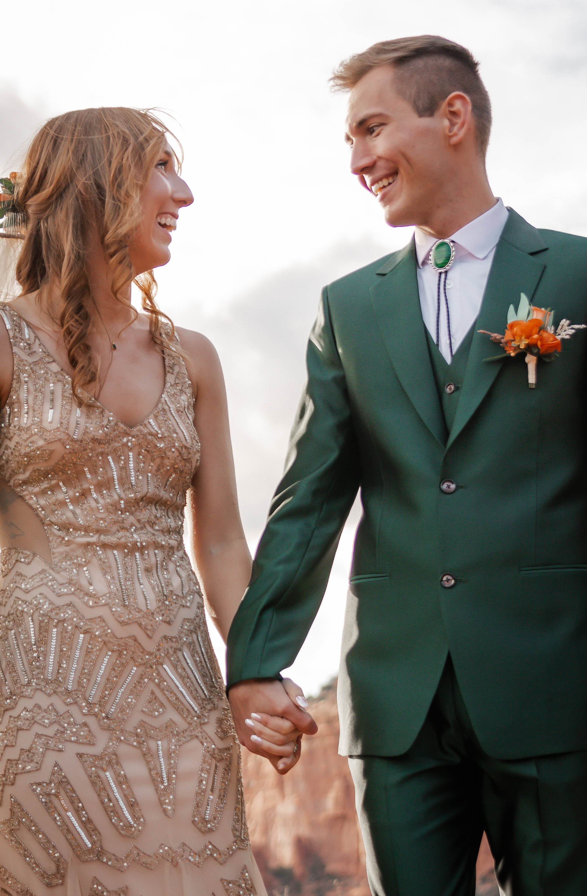 A bride and groom holding hands and smiling, outdoors in a cloudy sky setting.