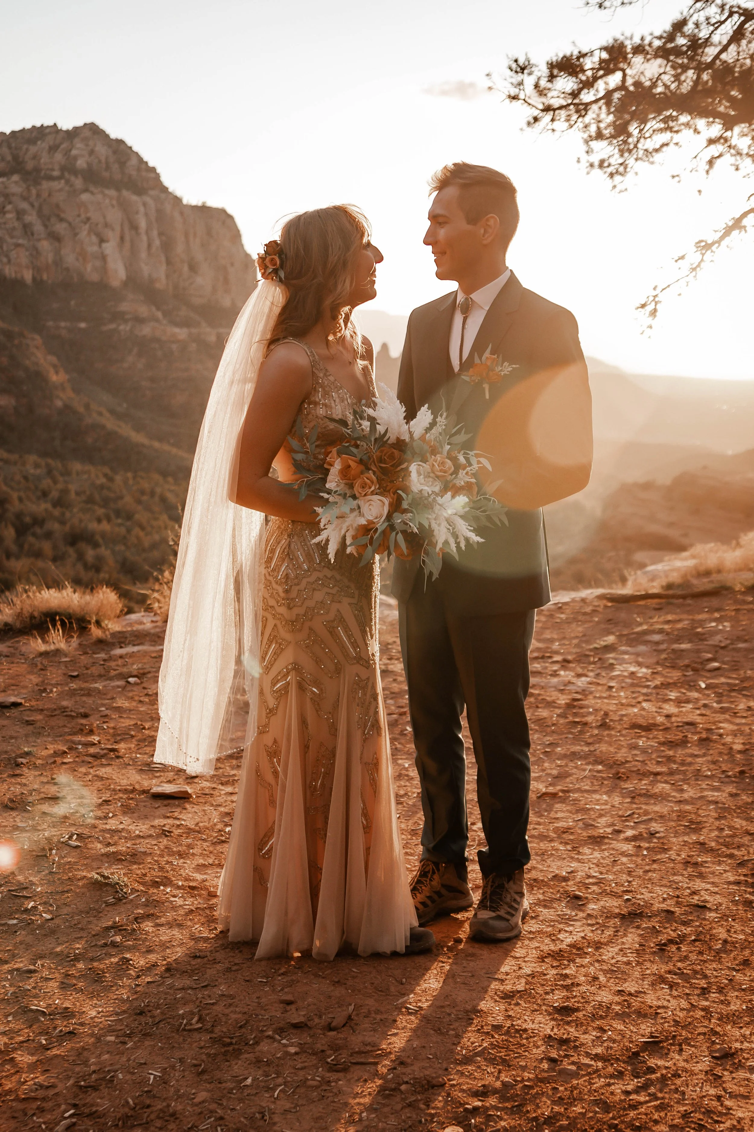 A bride and groom standing close together outdoors during sunset, with mountains and a tree in the background, celebrating their wedding.