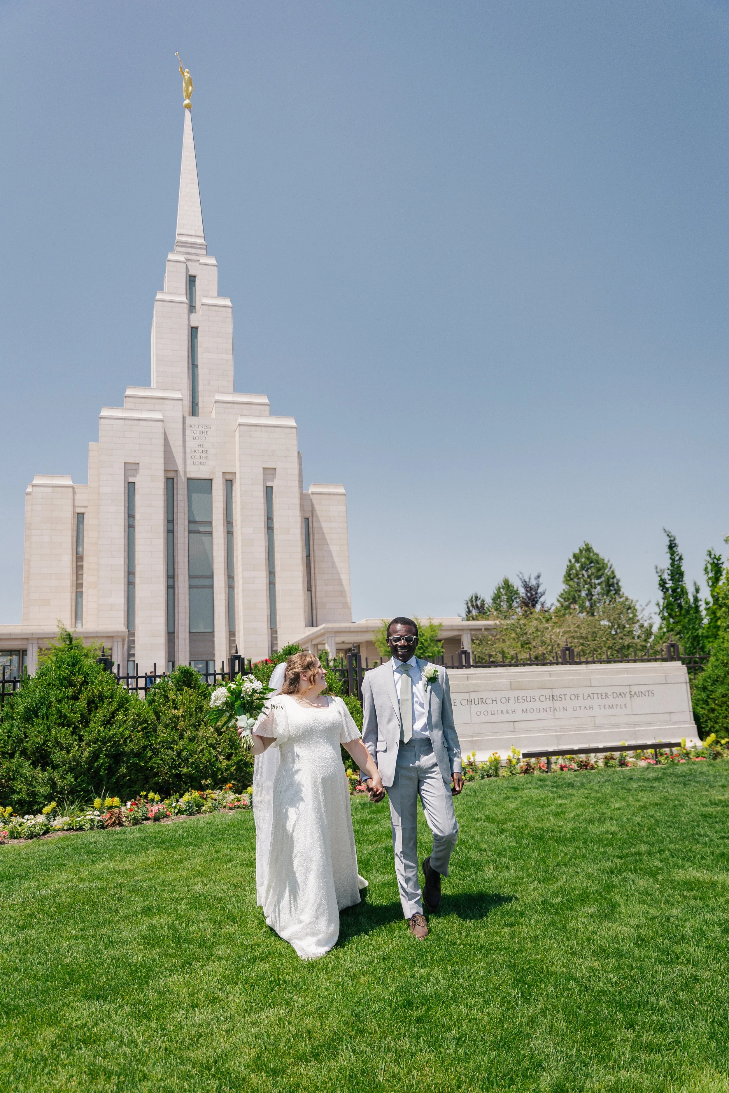 A newlywed couple holding hands and smiling while walking on a grassy area in front of the oquirrh mountain temple with green bushes, flowers, on a sunny day.
