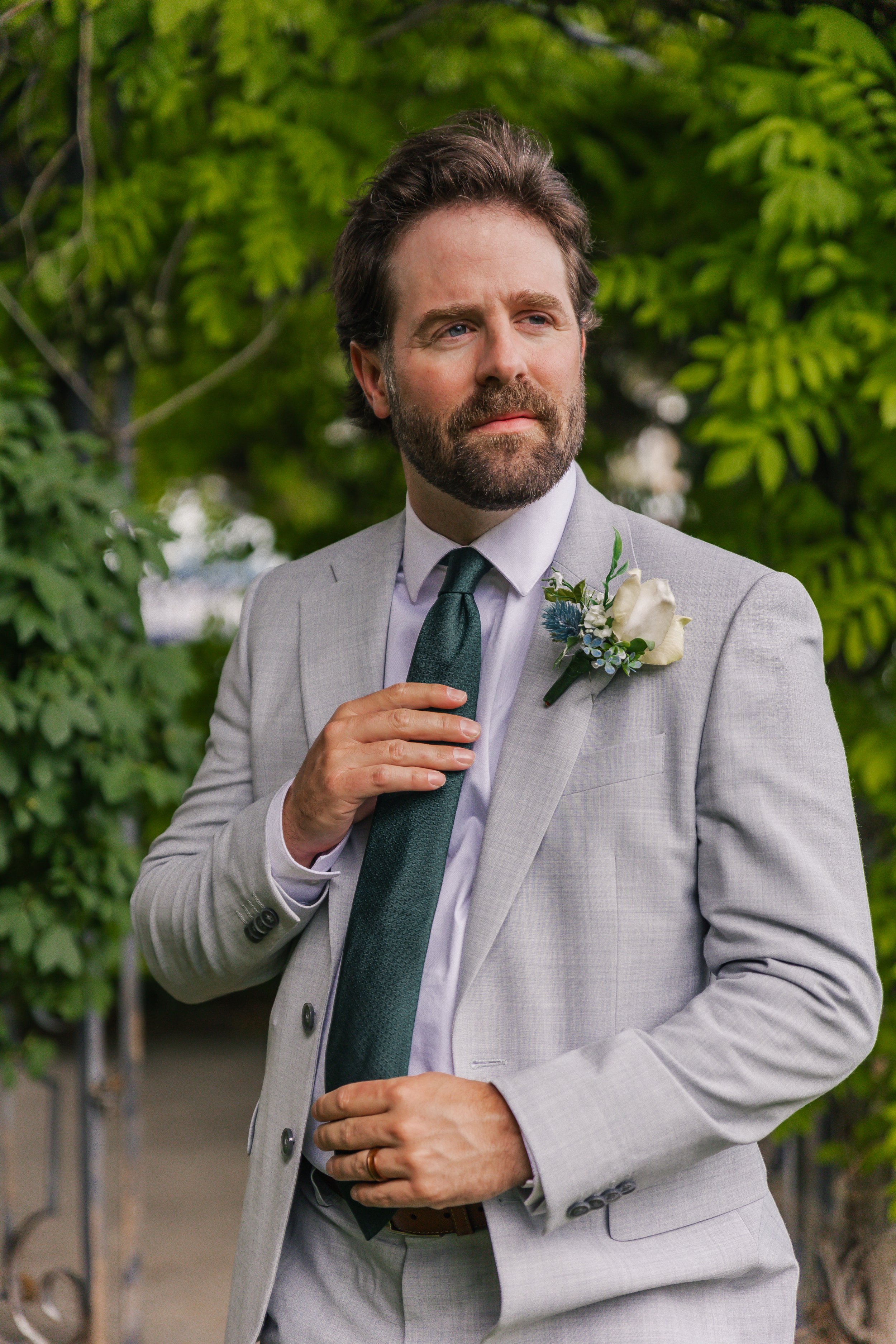 A man with a beard and brown hair, dressed in a light gray suit, white shirt, and dark green tie, stands outdoors among green trees. He has a white boutonniere with green leaves and small blue flowers on his left lapel. He is touching his chest with 
