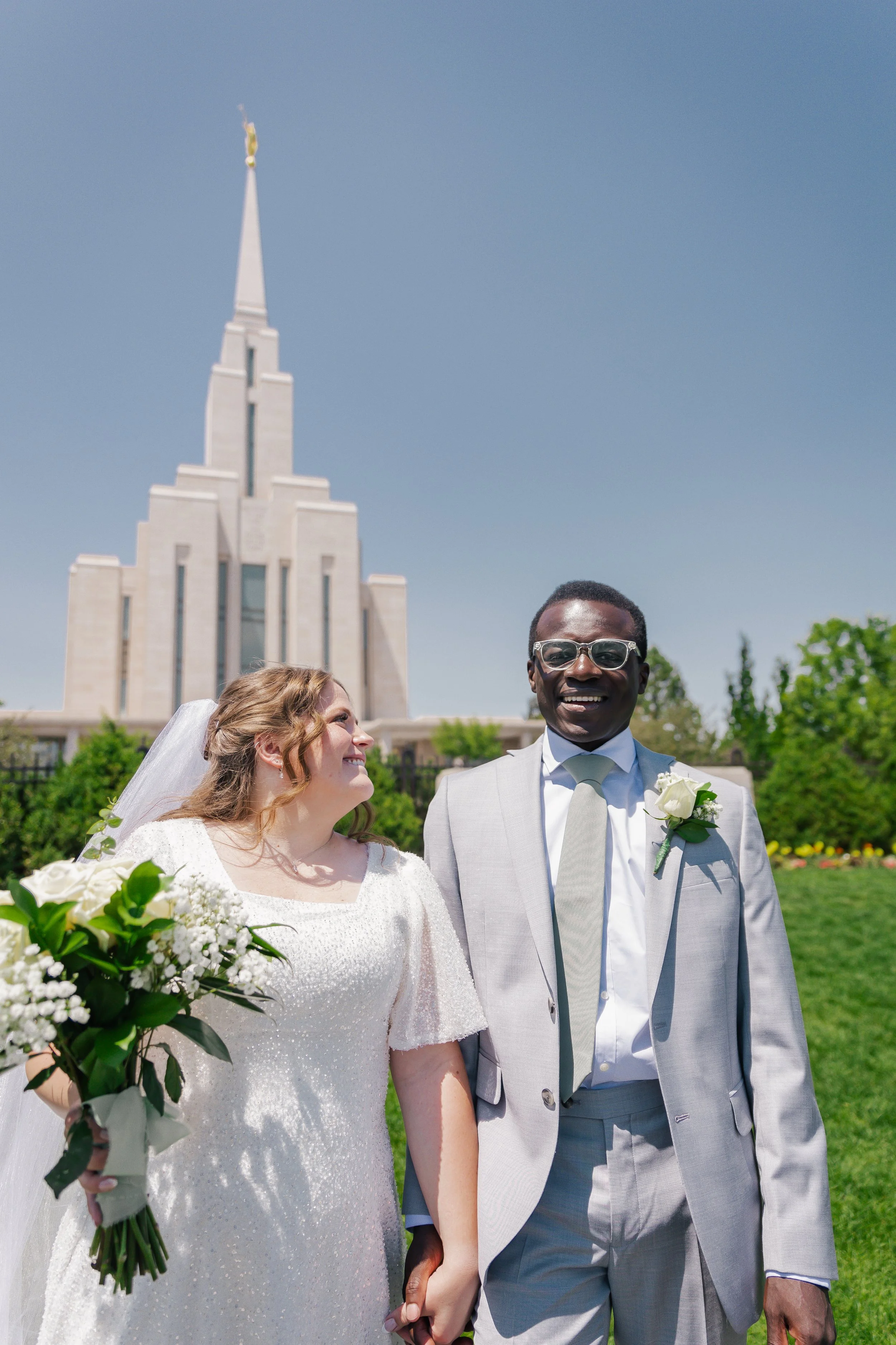 A newlywed couple in wedding attire holding hands outdoors on a sunny day; the bride carries a bouquet of white flowers and the groom wears glasses and a boutonnière, with oquirrh mountain temple in the background.