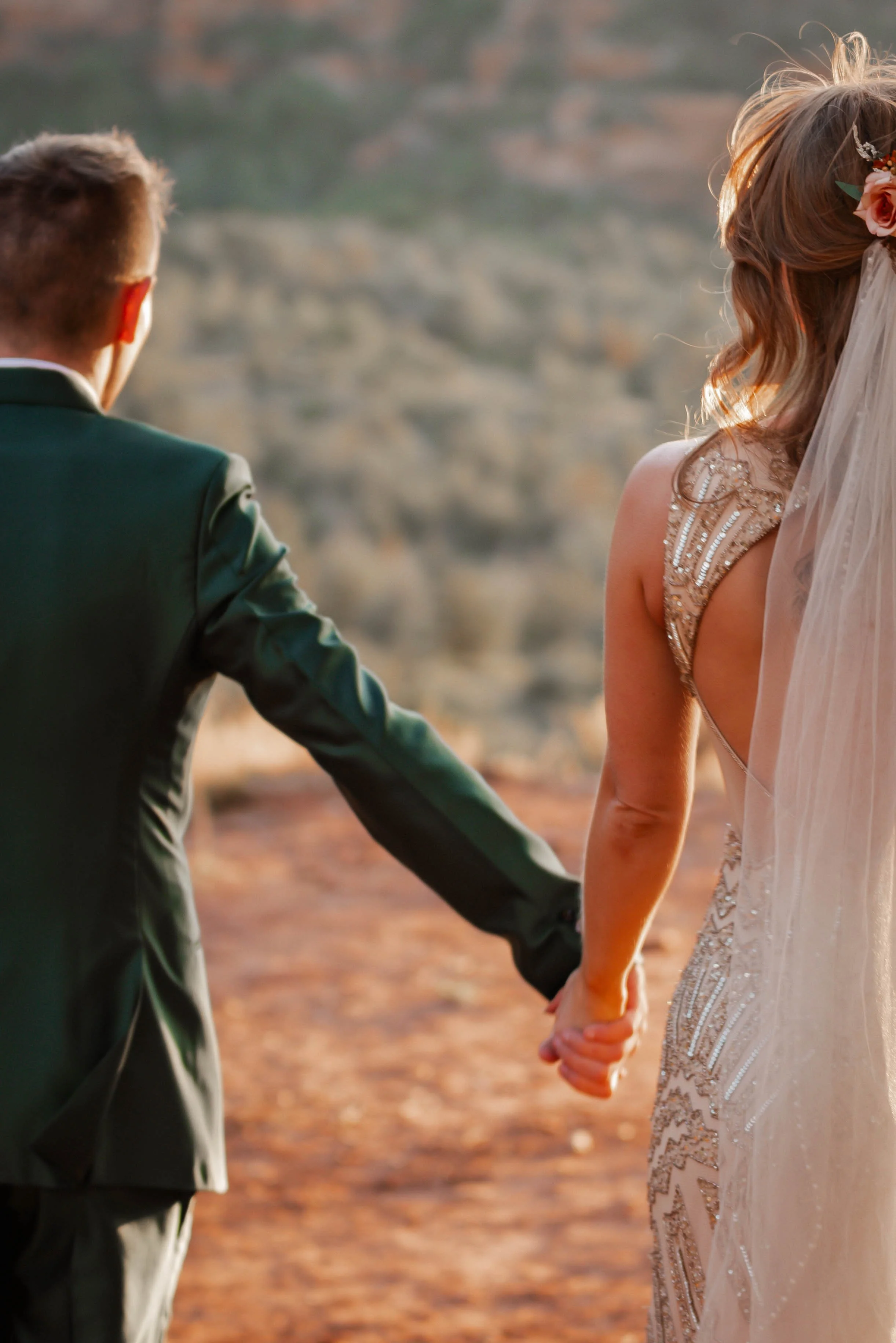 A couple holding hands outdoors during sunset, with a rocky landscape in the background. The woman is wearing an elegant, beaded wedding dress and a veil, while the man is dressed in a dark suit.