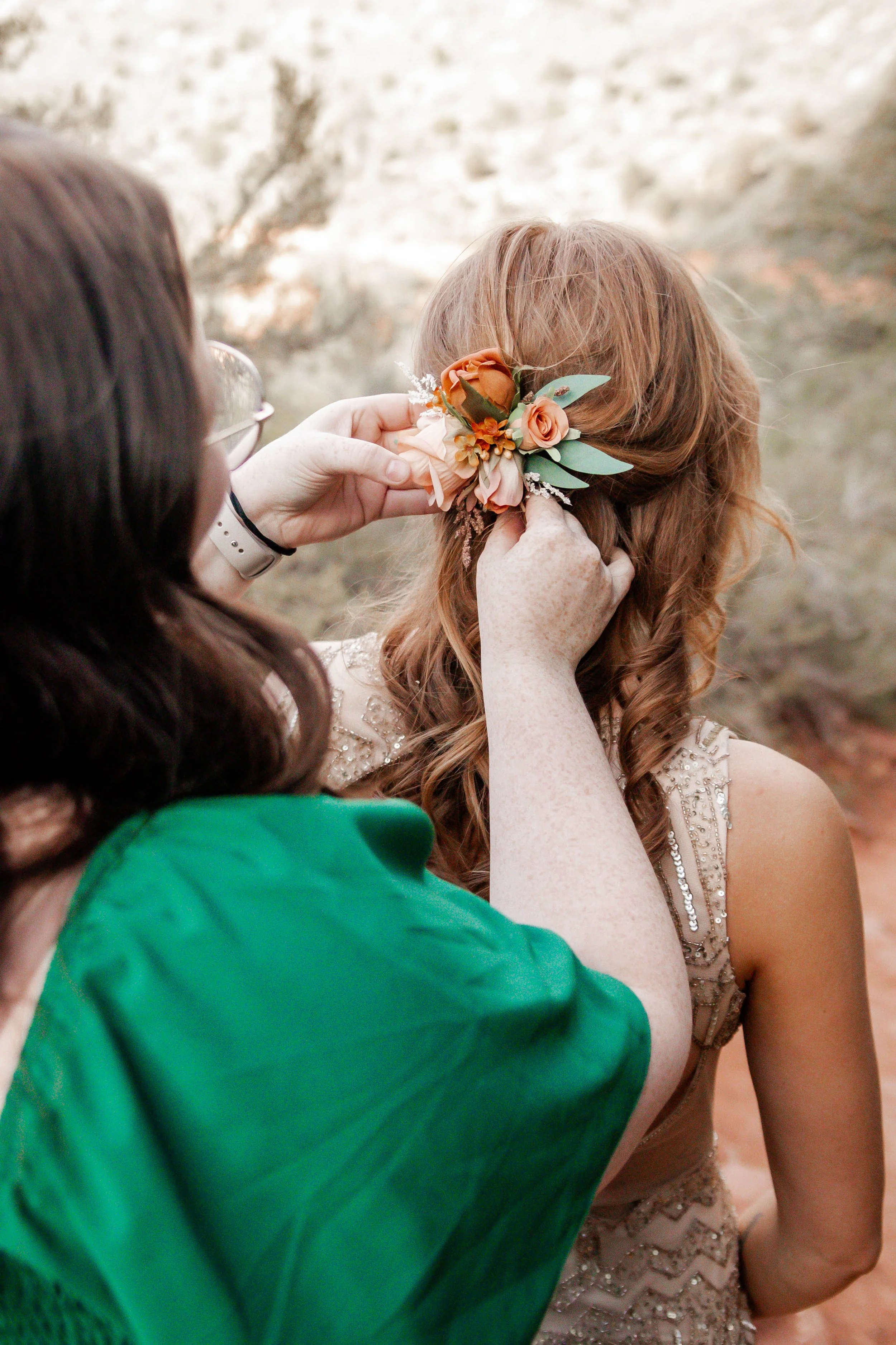 A woman with long brown hair is helping a red-haired woman put on a floral hair accessory in a natural outdoor setting.