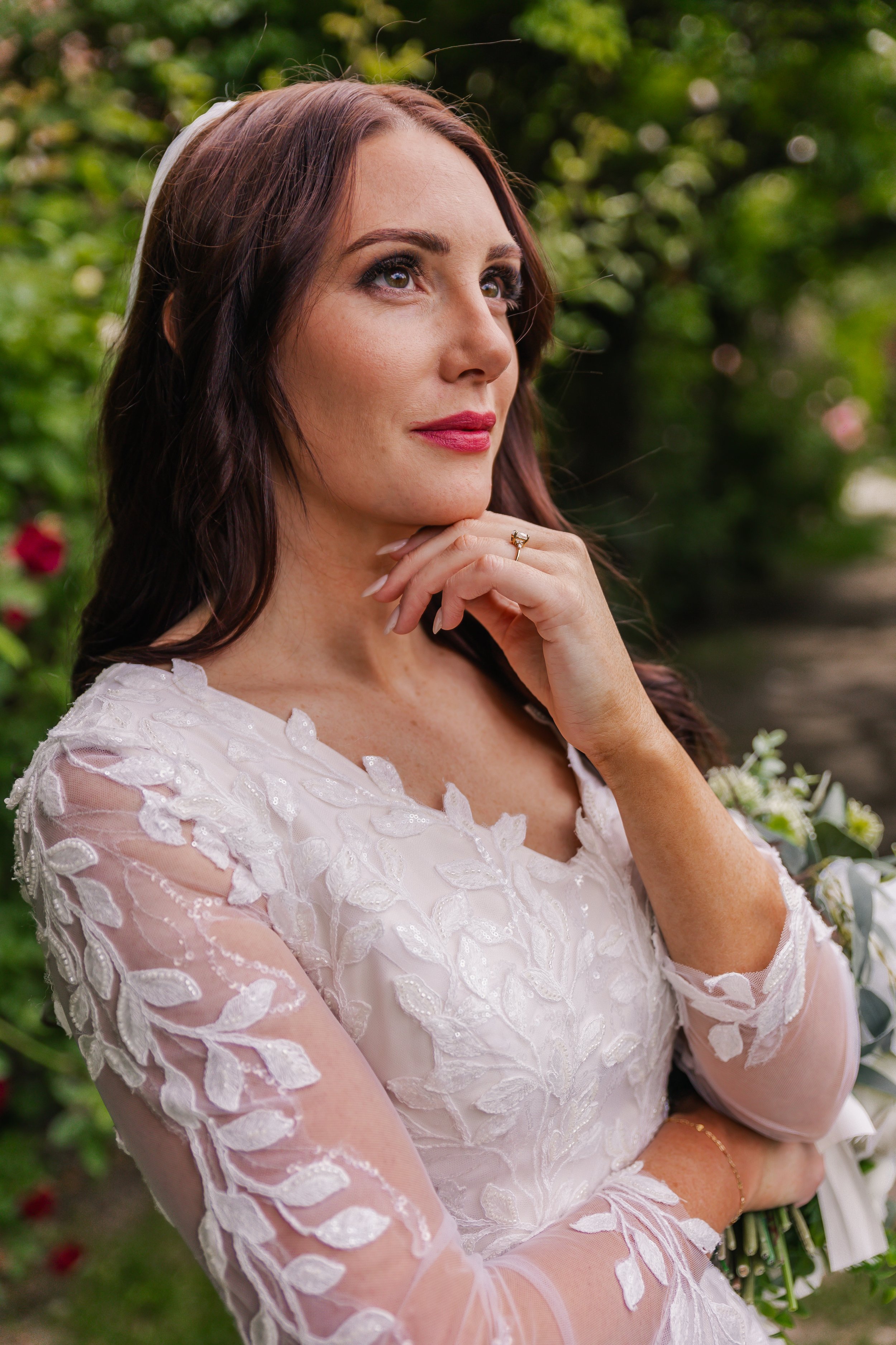 A woman in a white lace wedding dress outdoors with greenery and flowers in the background, looking thoughtfully to the side, hand resting near her chin.