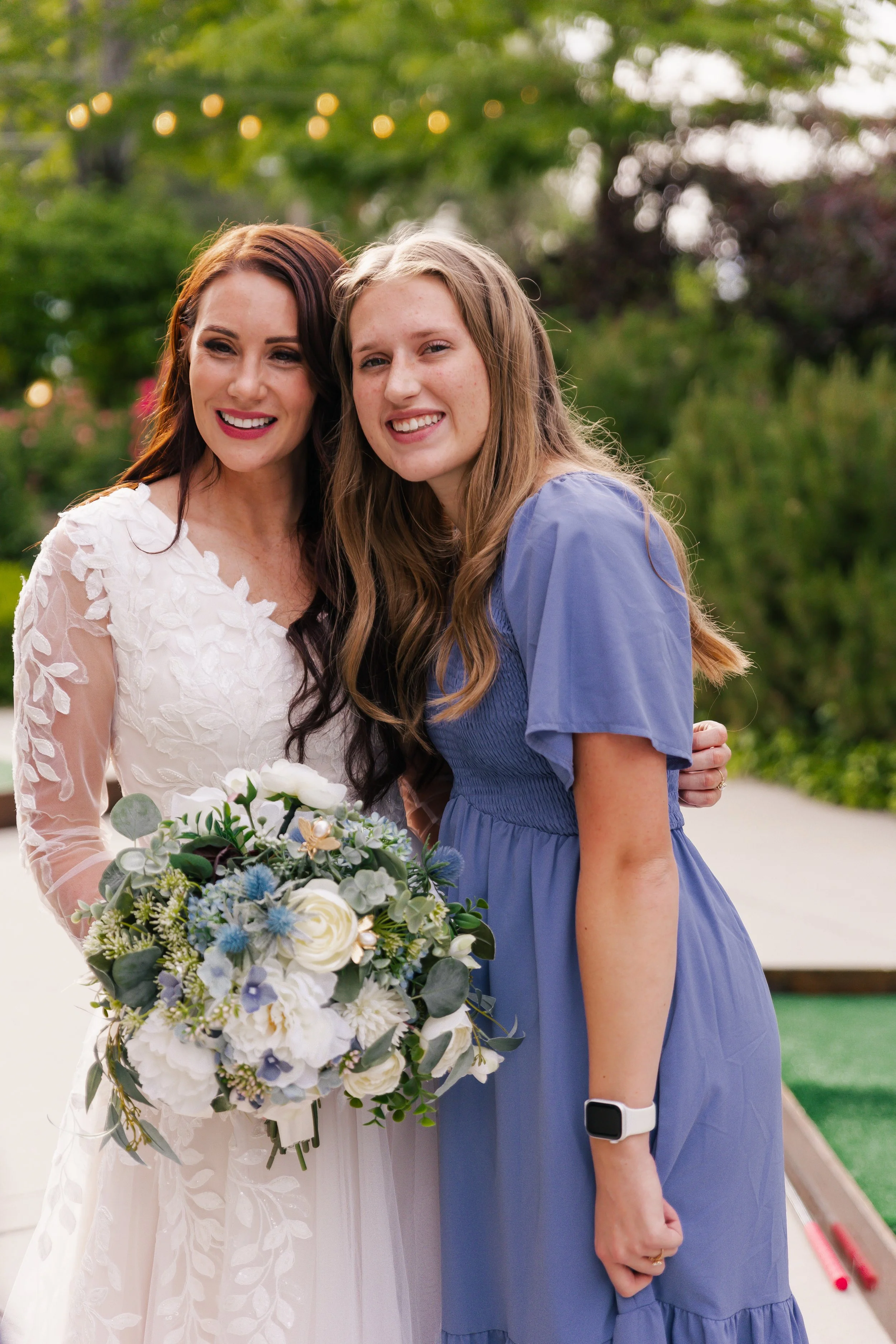 A bride in a white lace wedding dress holding a bouquet of white and blue flowers, standing next to a woman in a blue dress, outdoors with green trees and string lights in the background.
