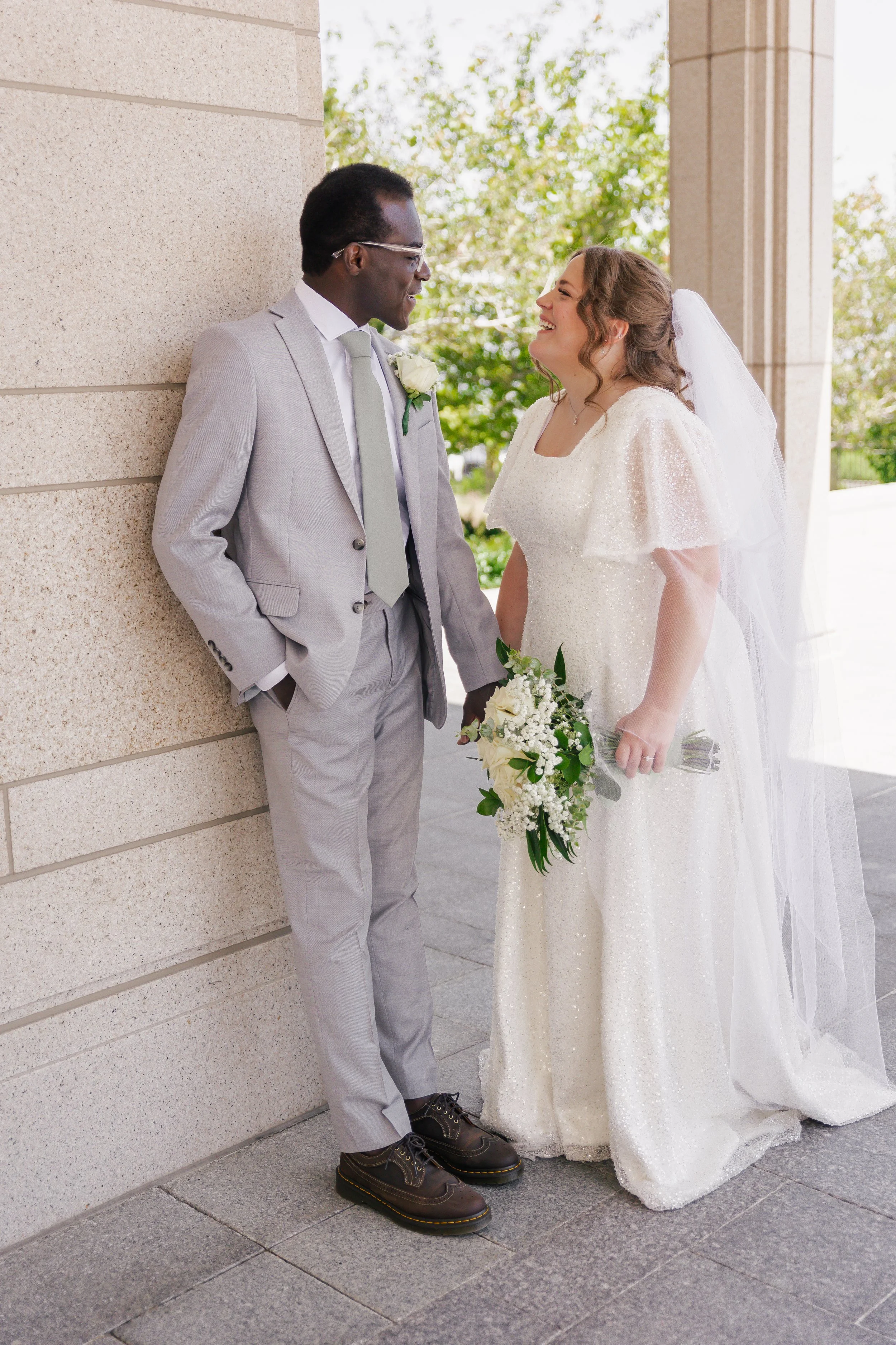 A bride and groom stand together smiling outside a building, holding hands. The groom wears a light gray suit and glasses, and the bride wears a white wedding dress with a veil, holding a bouquet of white flowers, with leafy accents.