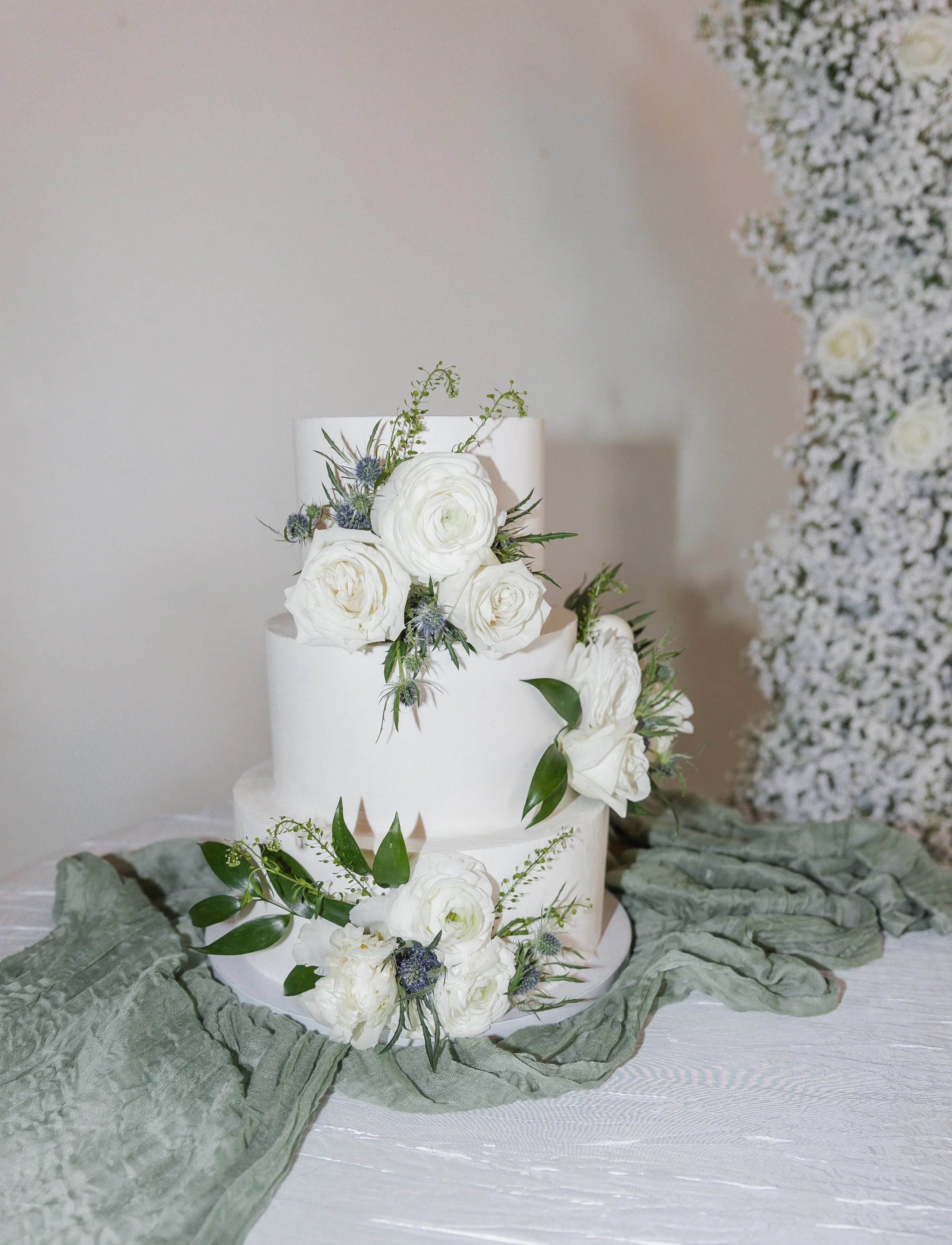 Three-tiered white wedding cake decorated with white roses, greenery, and thistle, placed on a table with a green cloth and a background of white floral decorations.