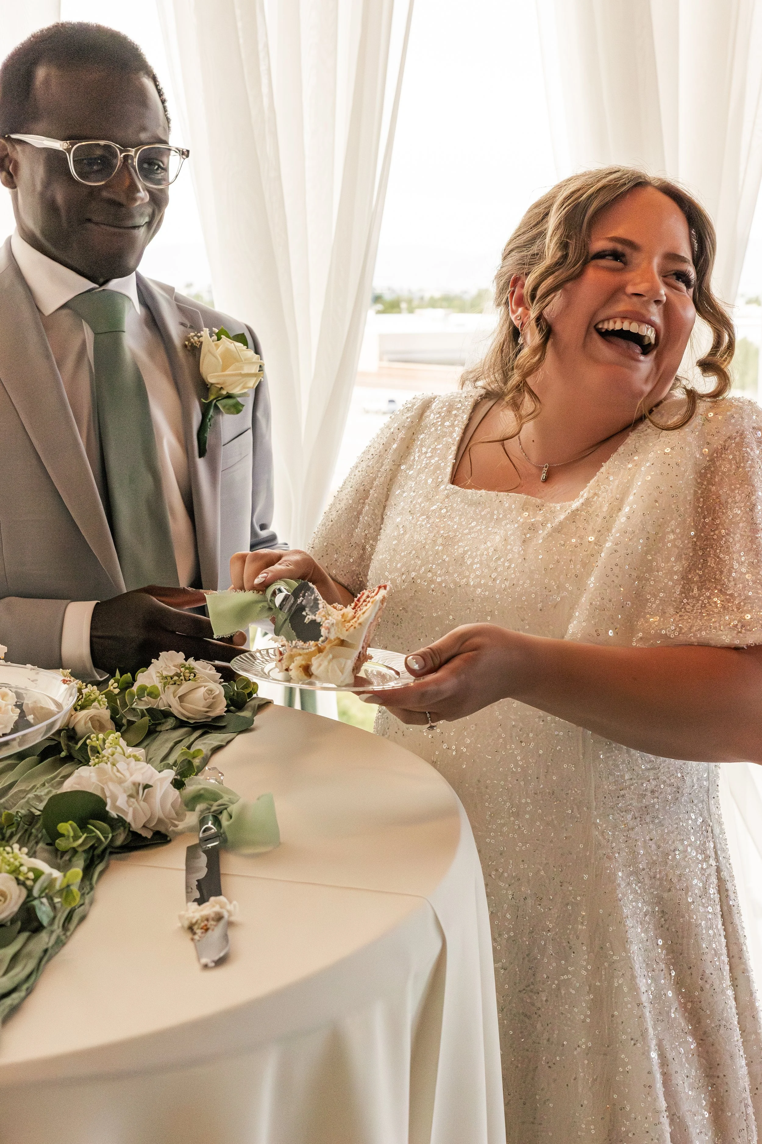 A bride and groom exchanging slices of cake during their wedding reception, smiling and laughing.