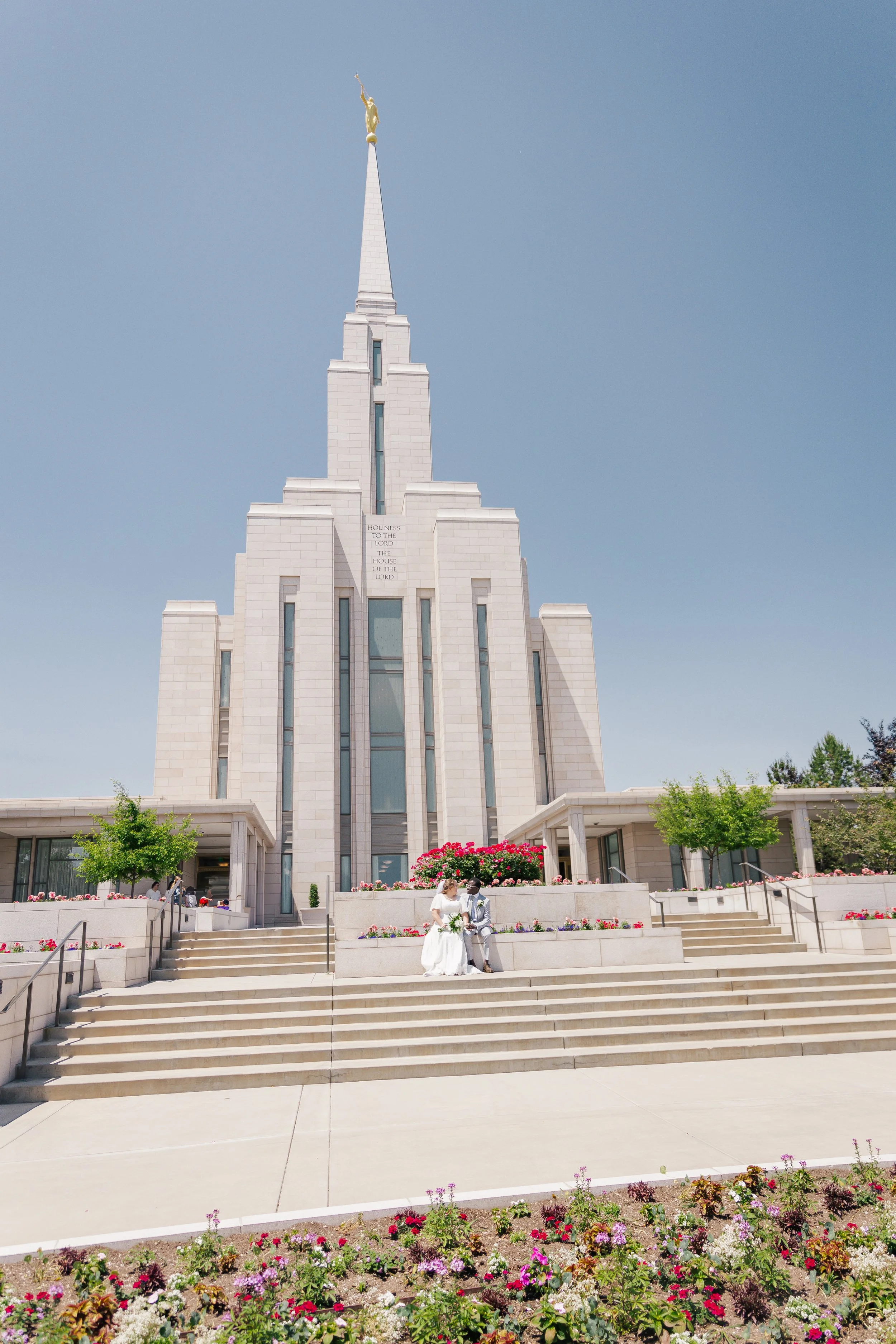 A wedding couple sitting on steps in front the oquirrh mountain temple topped with a golden angel statue, and colorful flowers in the foreground.