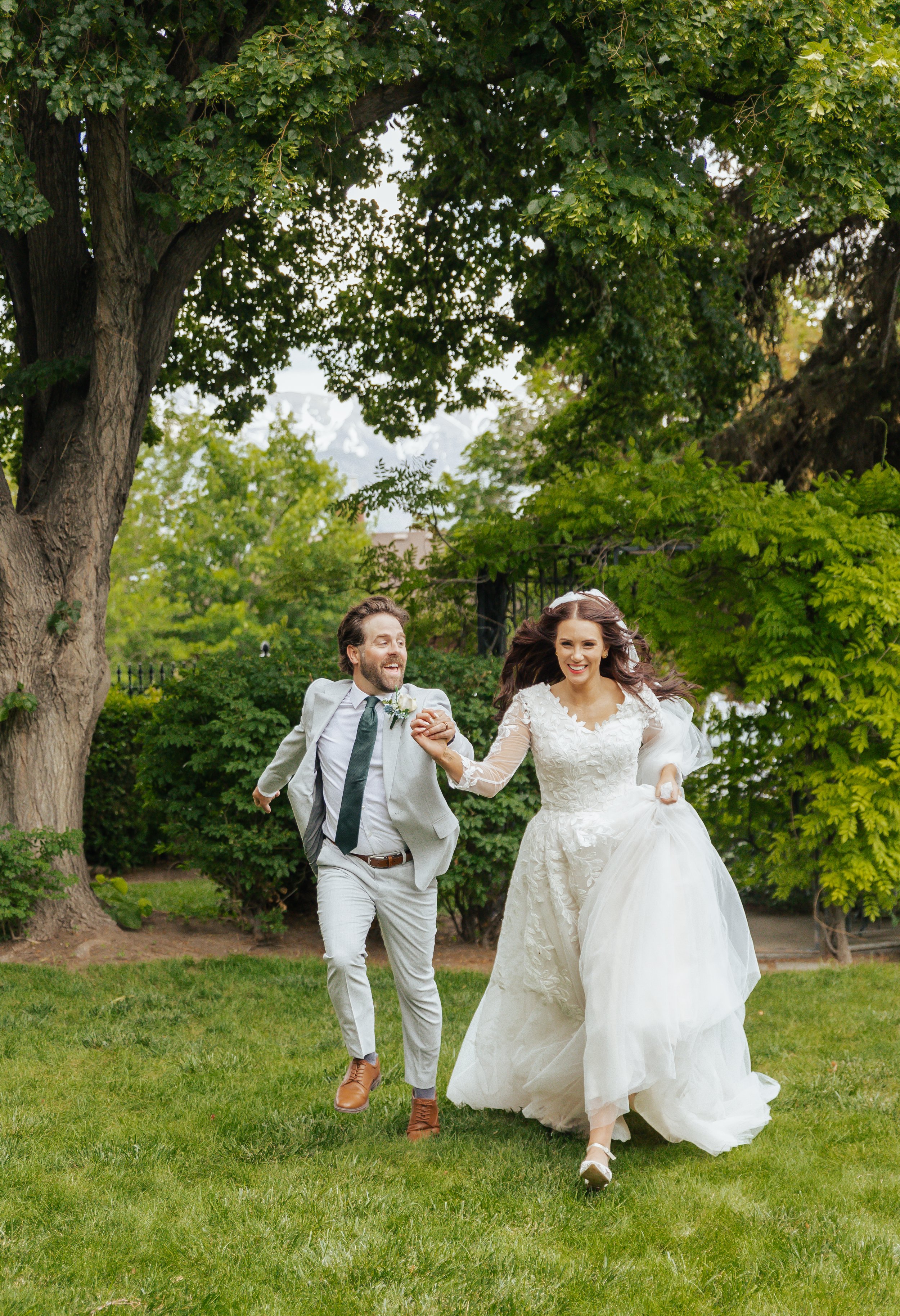 A bride and groom running hand in hand on a grassy lawn with trees in the background.
