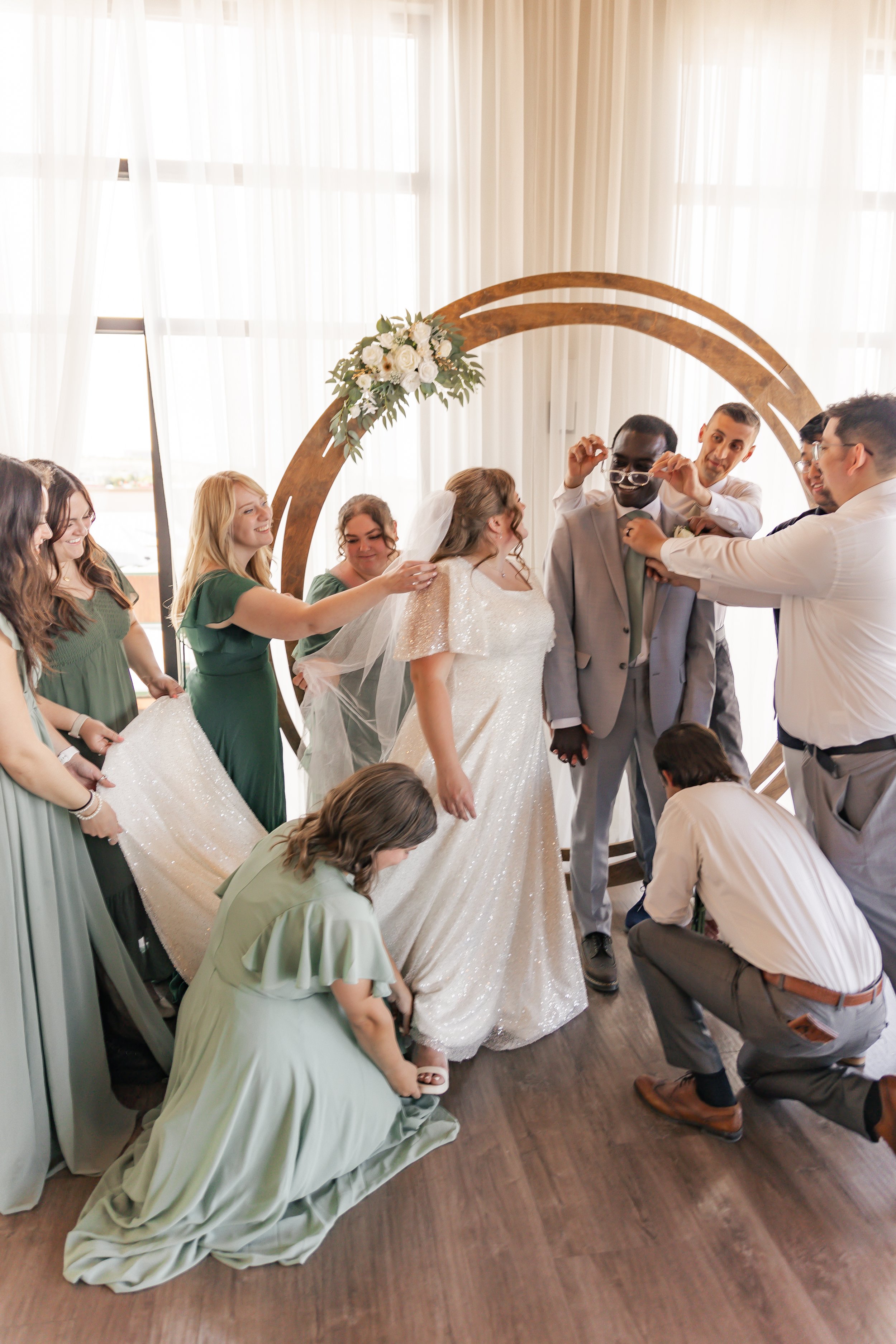 A bride and groom are surrounded by friends during their wedding ceremony. The bride wears a white, sparkling wedding dress and veil, while the guests, dressed in green and gray, assist with their attire. A decorative arch with white flowers is in th