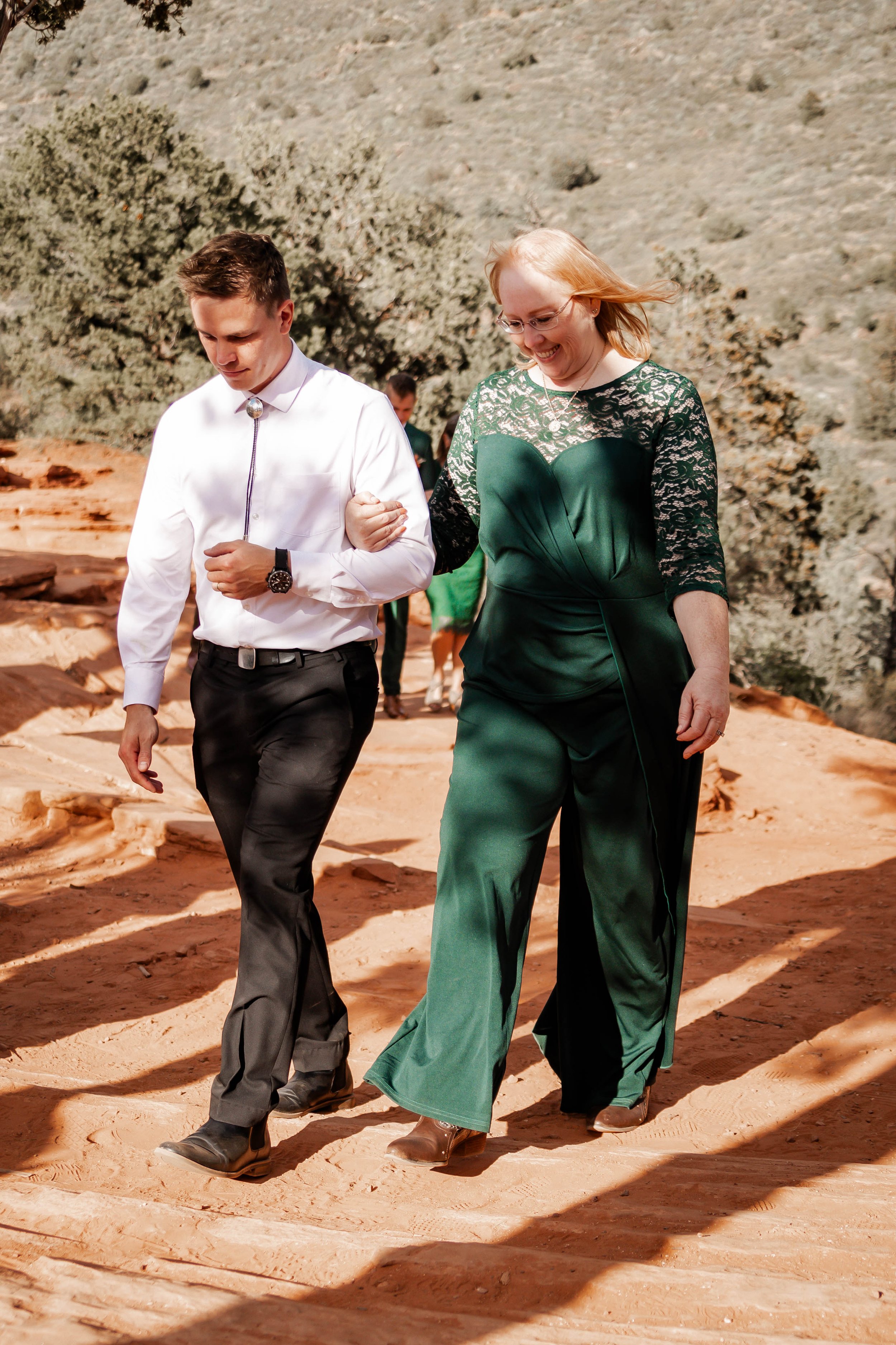 A woman and a young man walk together on a dirt trail in a desert landscape, with the woman supporting the man by holding his arm. The woman wears a green dress with lace details, and the young man wears a white shirt and black pants.