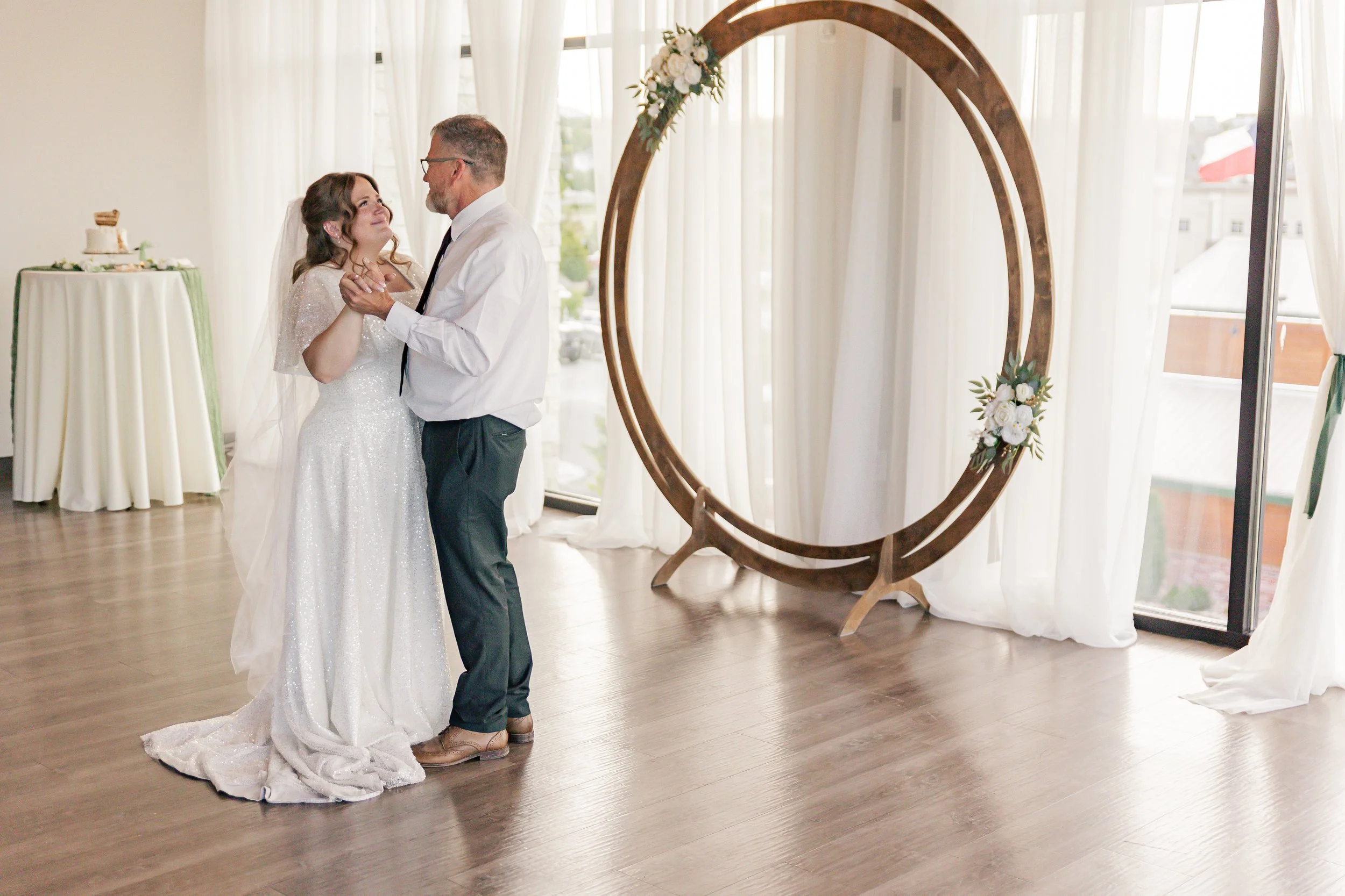 A bride and an older man, possibly her father, dance together at a wedding reception. The bride wears a white wedding gown and veil, and they hold each other while smiling. In the background, there is a circular floral decoration, large windows with 