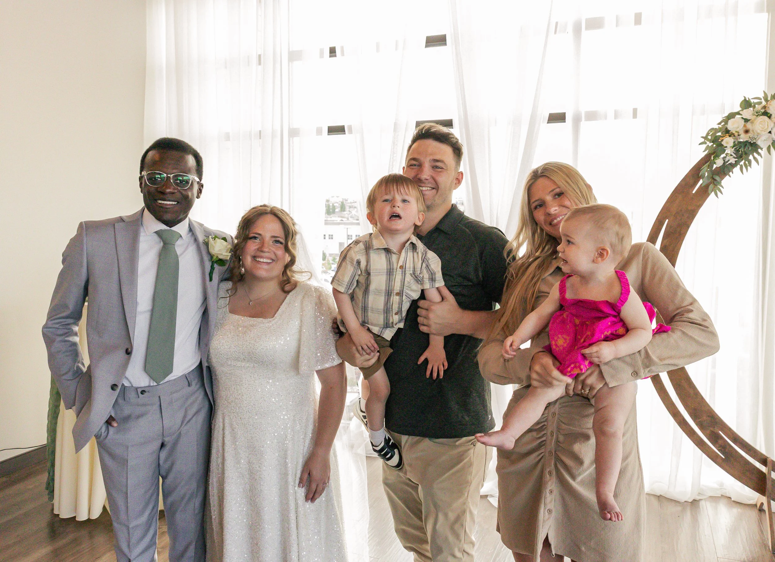 Group of six people, including two children, smiling at a gathering in a well-lit room with sheer curtains and a decorative crescent moon, during daytime.