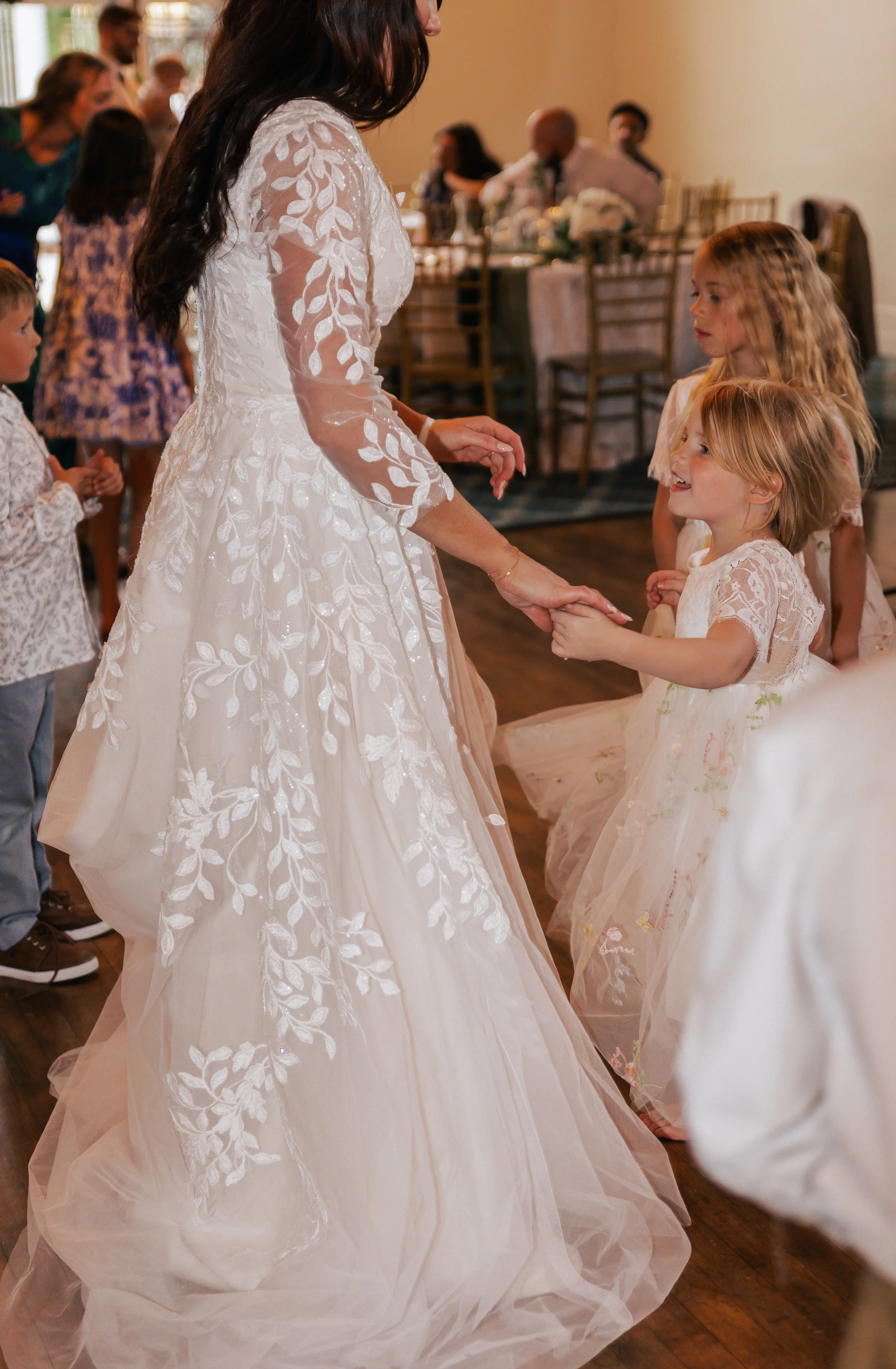 A woman in a white, wedding dress dances with a young girl in a light-colored dress at a wedding reception.